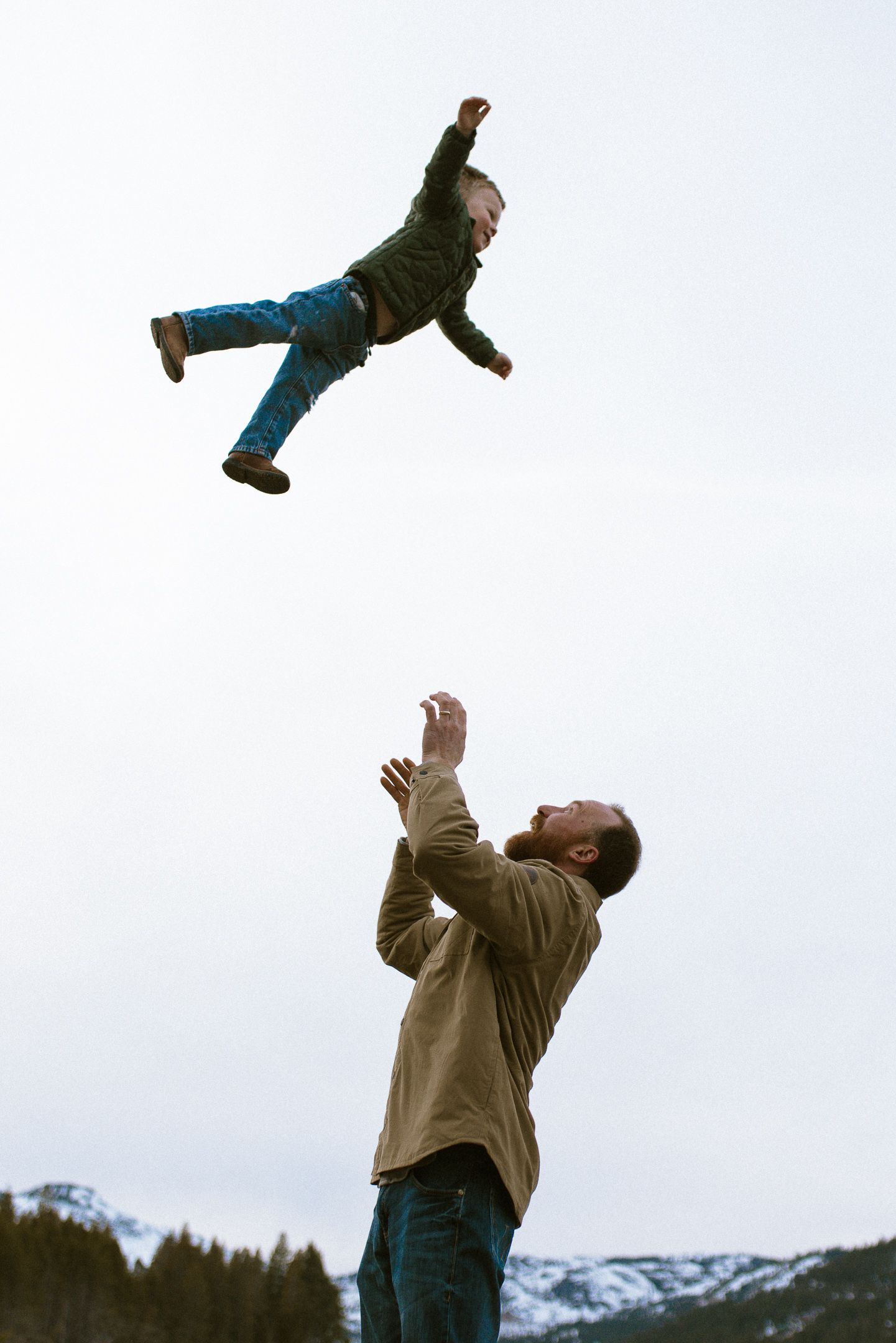 Father tossing his laughing child into the air against snowy mountain peaks, joyful outdoor family session