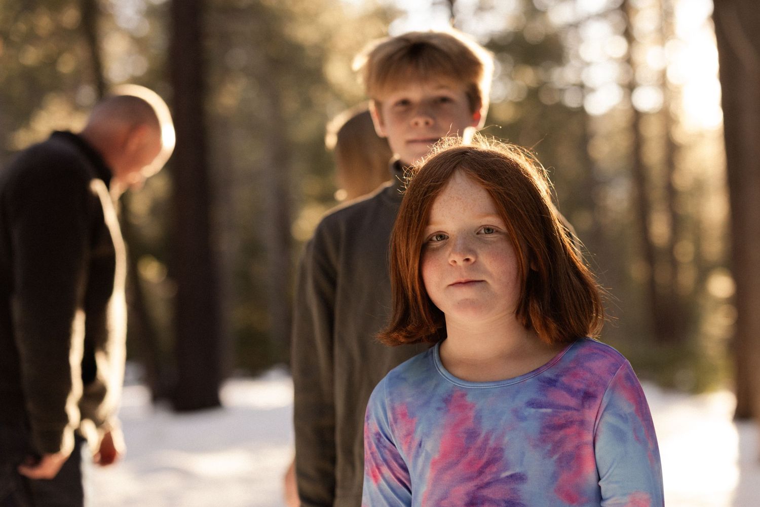 Young girl in a tie-dye shirt smiling at the camera with her family behind in a snowy sunlit forest, candid family session