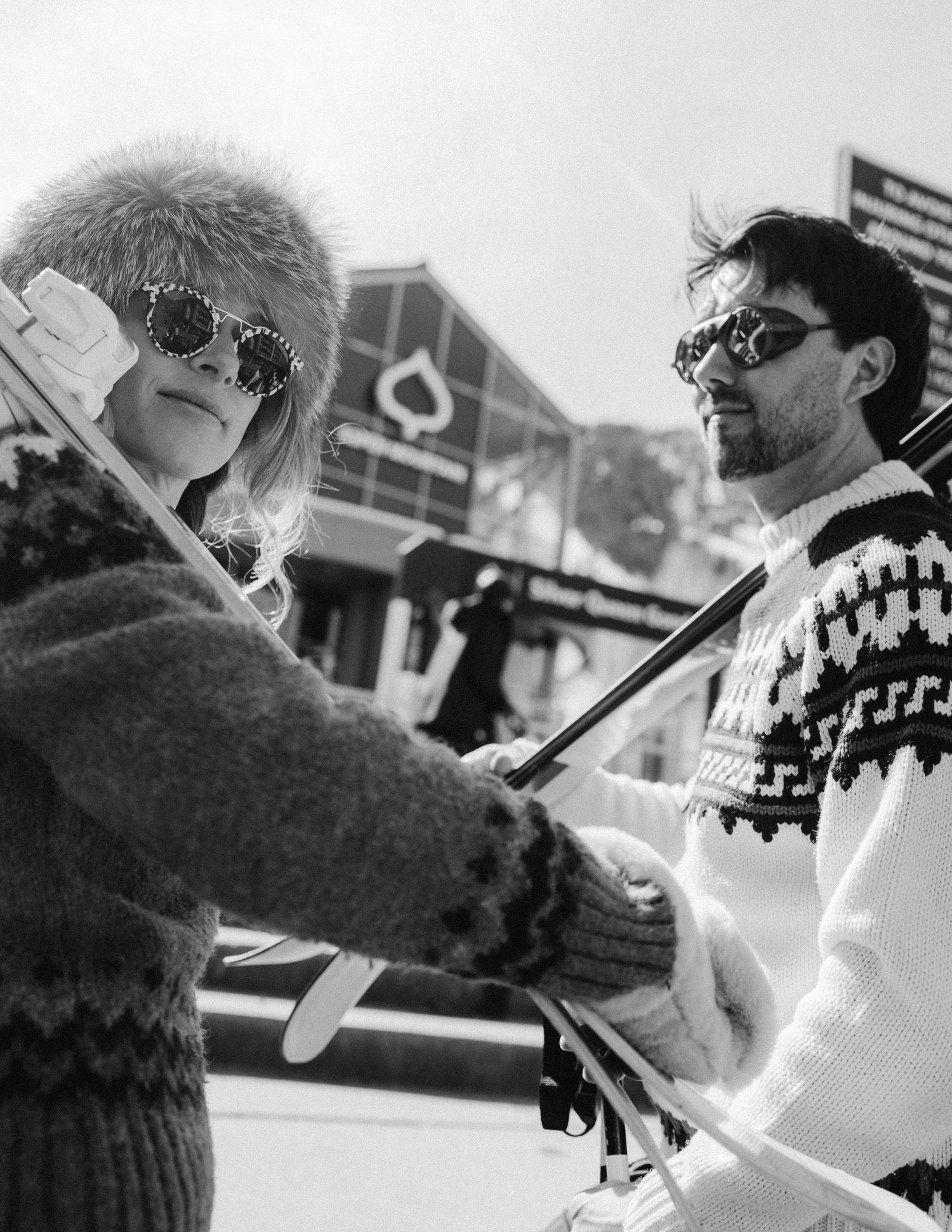Black-and-white photo of a stylish couple in retro ski gear holding poles at a snowy resort, playful winter engagement session