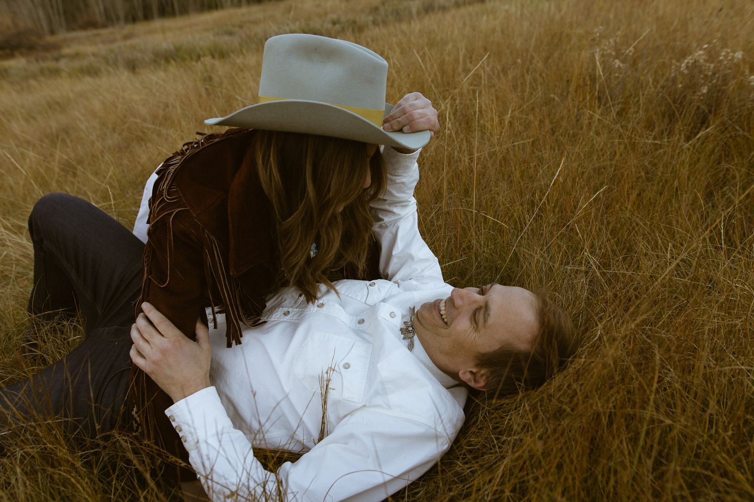 Couple lying in a golden field, one raising a hat playfully, joyful Western couples session