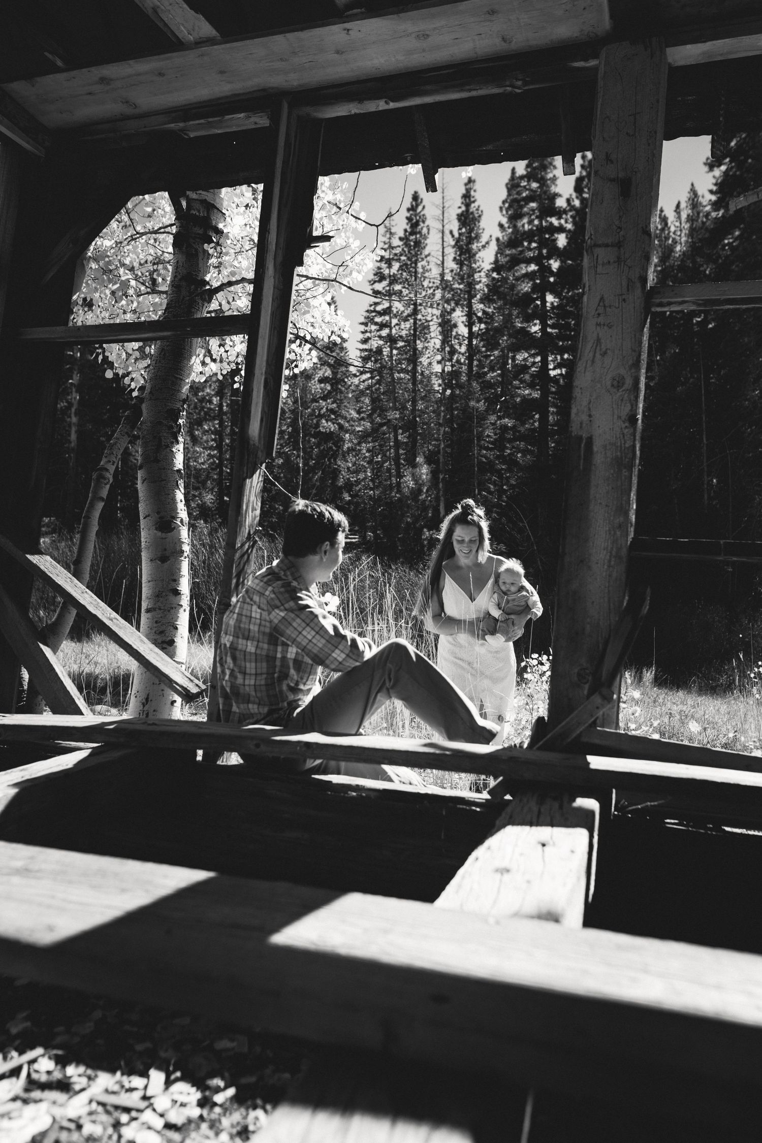 Black-and-white photo of a family relaxing on a cabin deck overlooking pines and a lake, candid outdoor family session