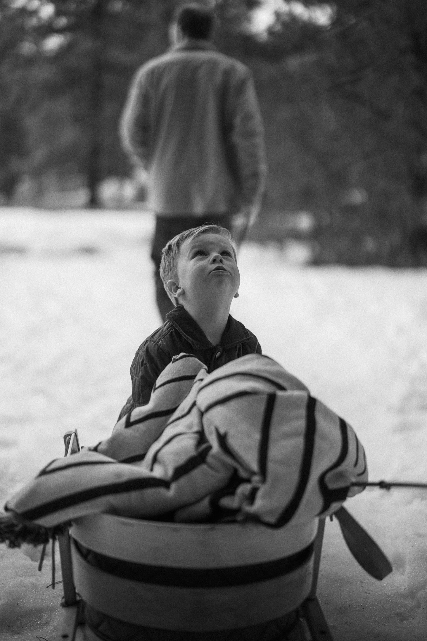 Black-and-white photo of a bundled-up boy gazing up from a sled in a snowy forest during a winter family session