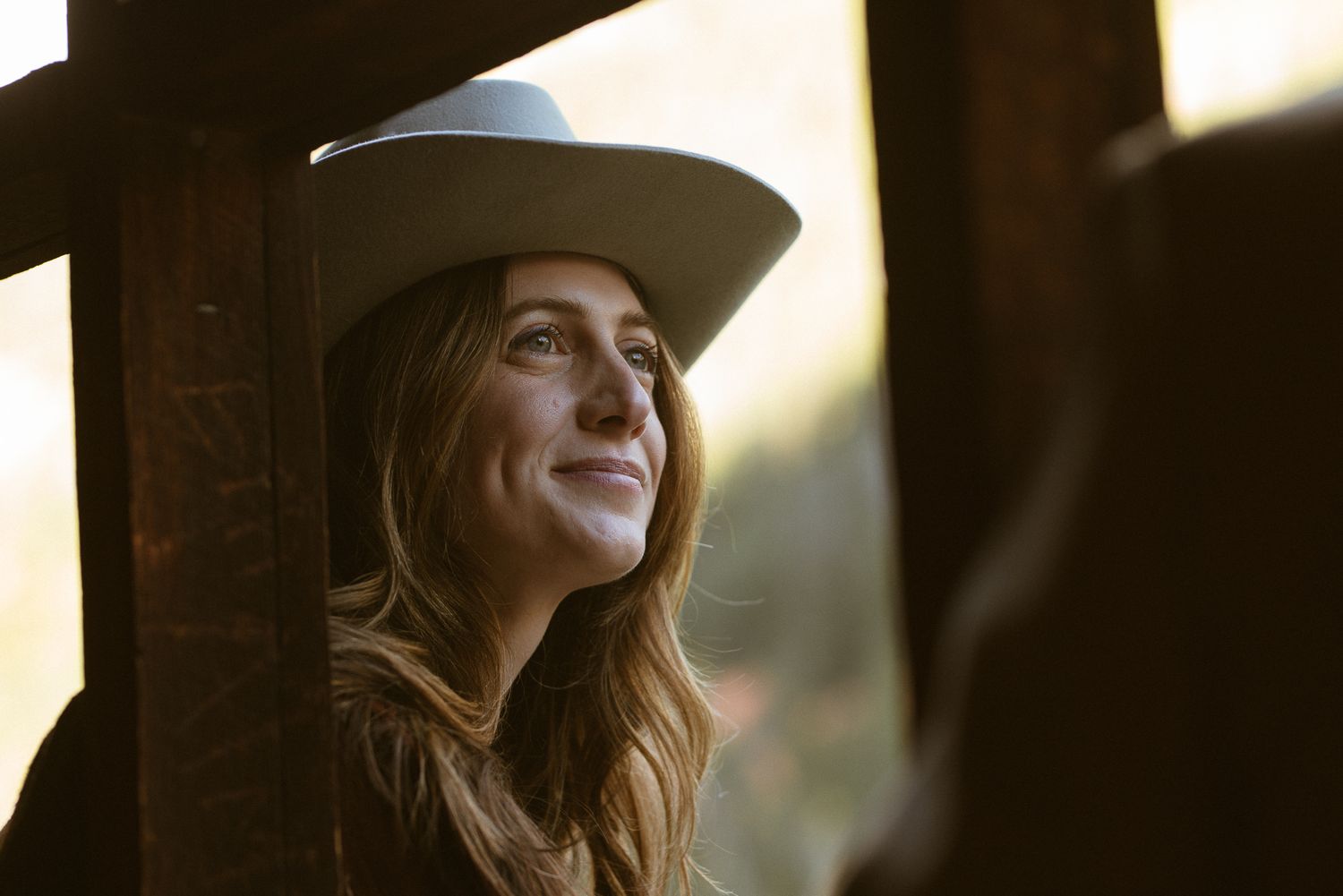 Woman in a wide-brim hat smiling against a wooden post at golden hour, warm lifestyle branding portrait