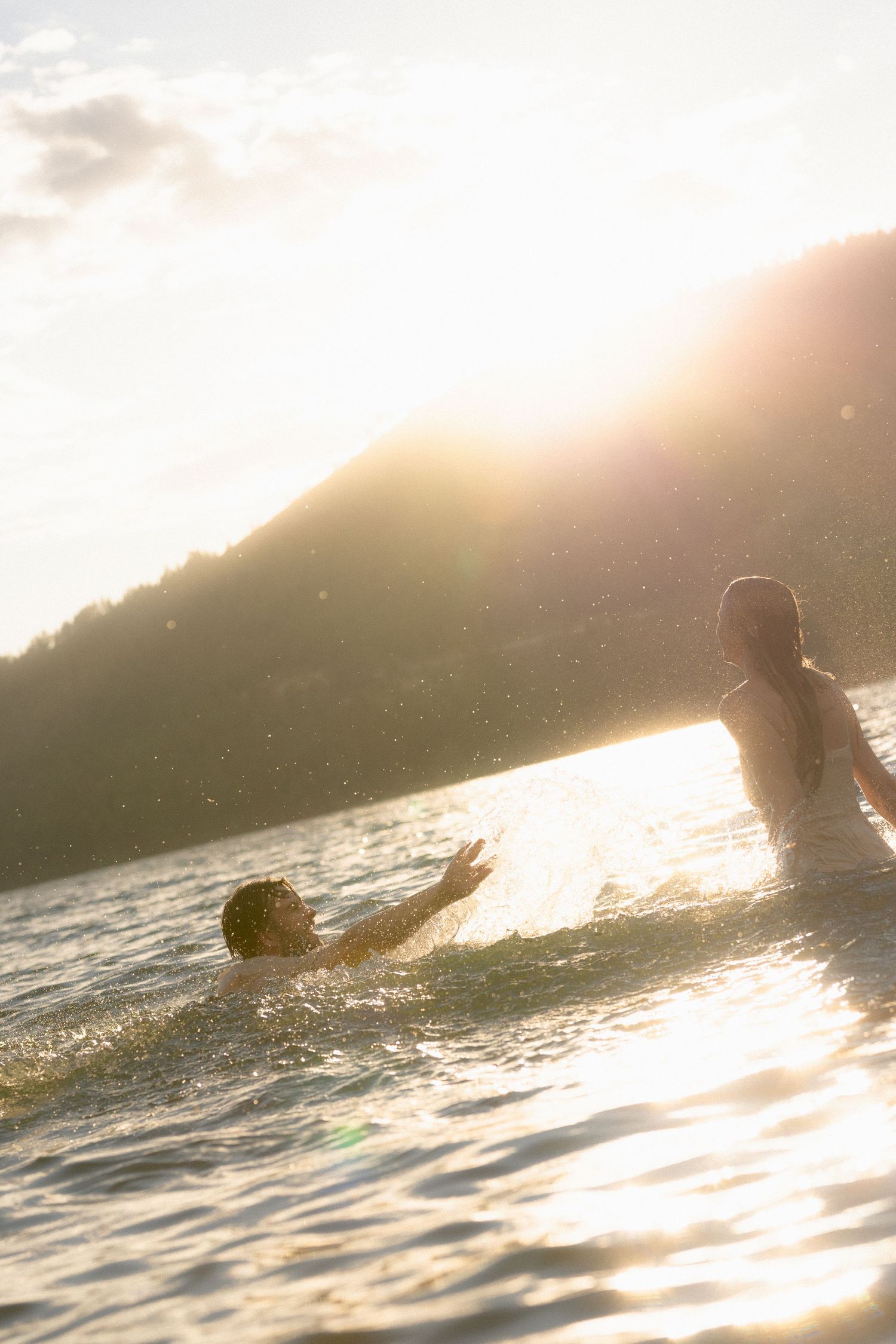 Couple wading into a mountain lake at sunset as the sun flares over the ridge, dreamy golden-hour story session