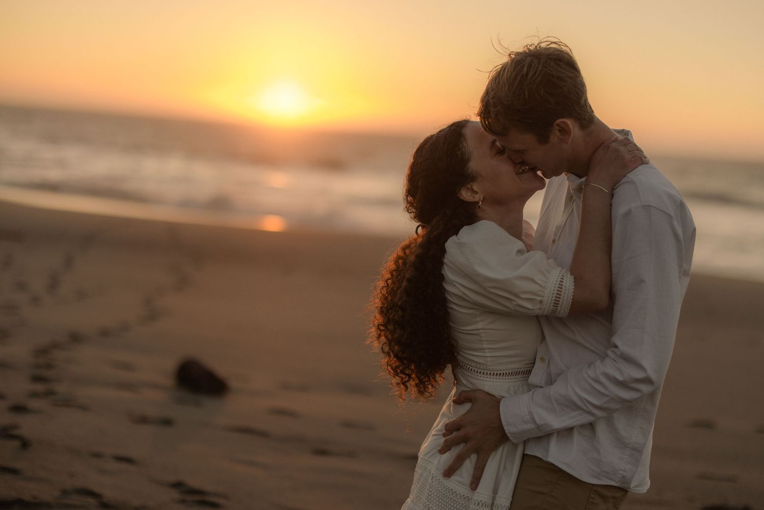 Couple kissing on the beach as the sun sets over the water, romantic coastal engagement session
