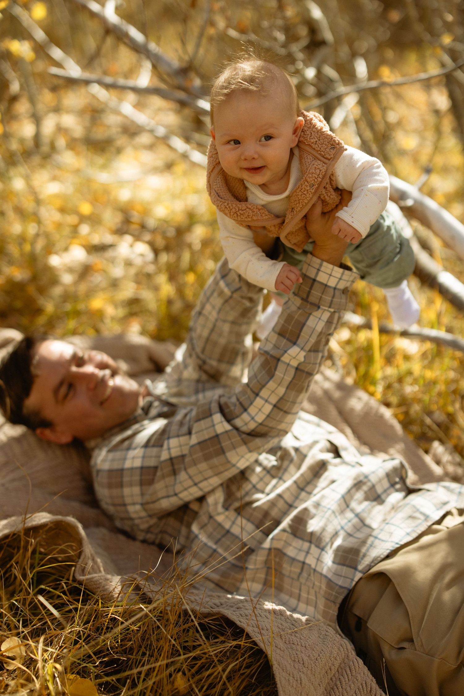 Father lying in golden grass lifting his smiling baby into the air, joyful autumn family session