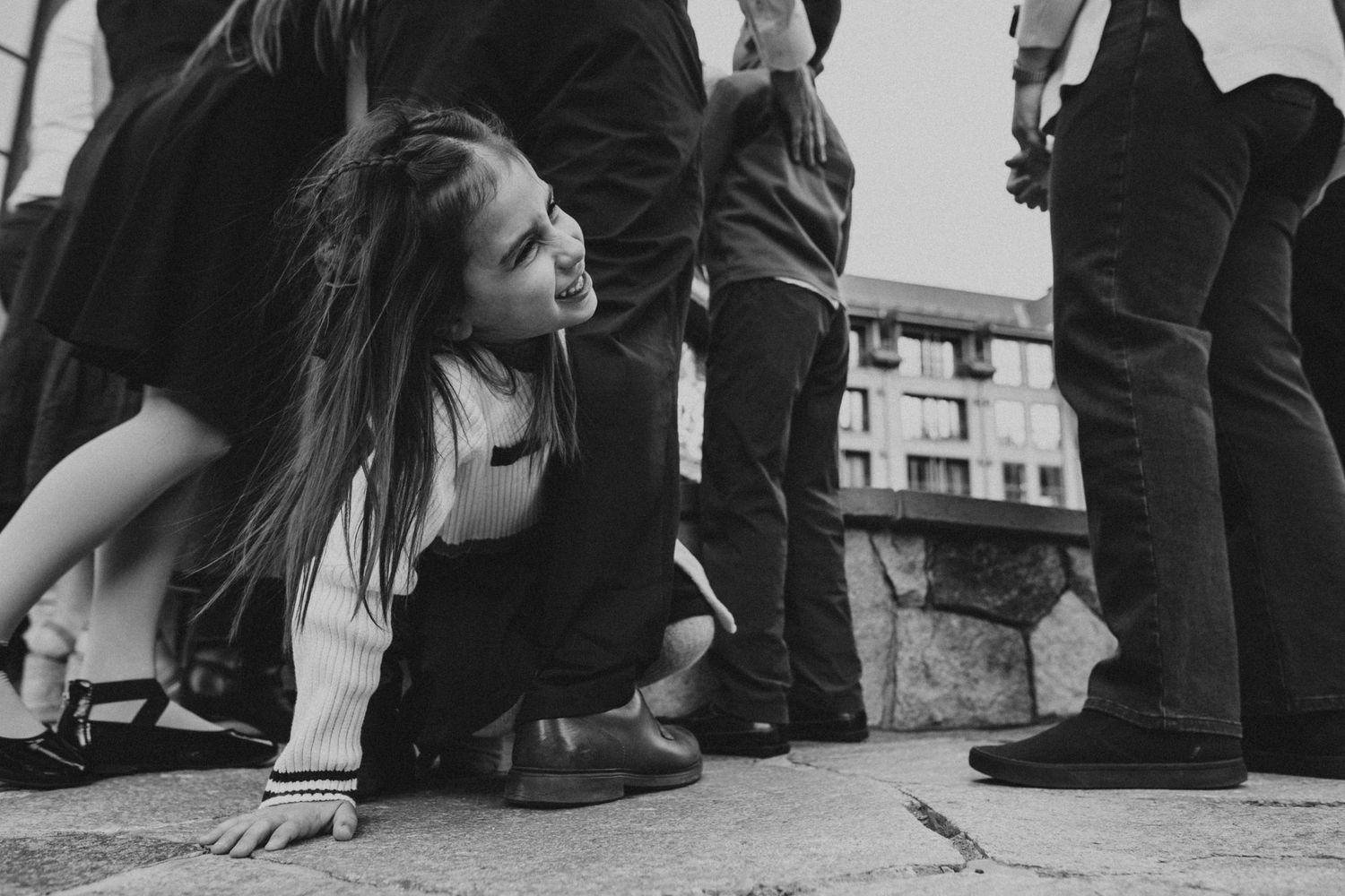 Black-and-white photo of a young girl crouching playfully among wedding guests, candid documentary moment