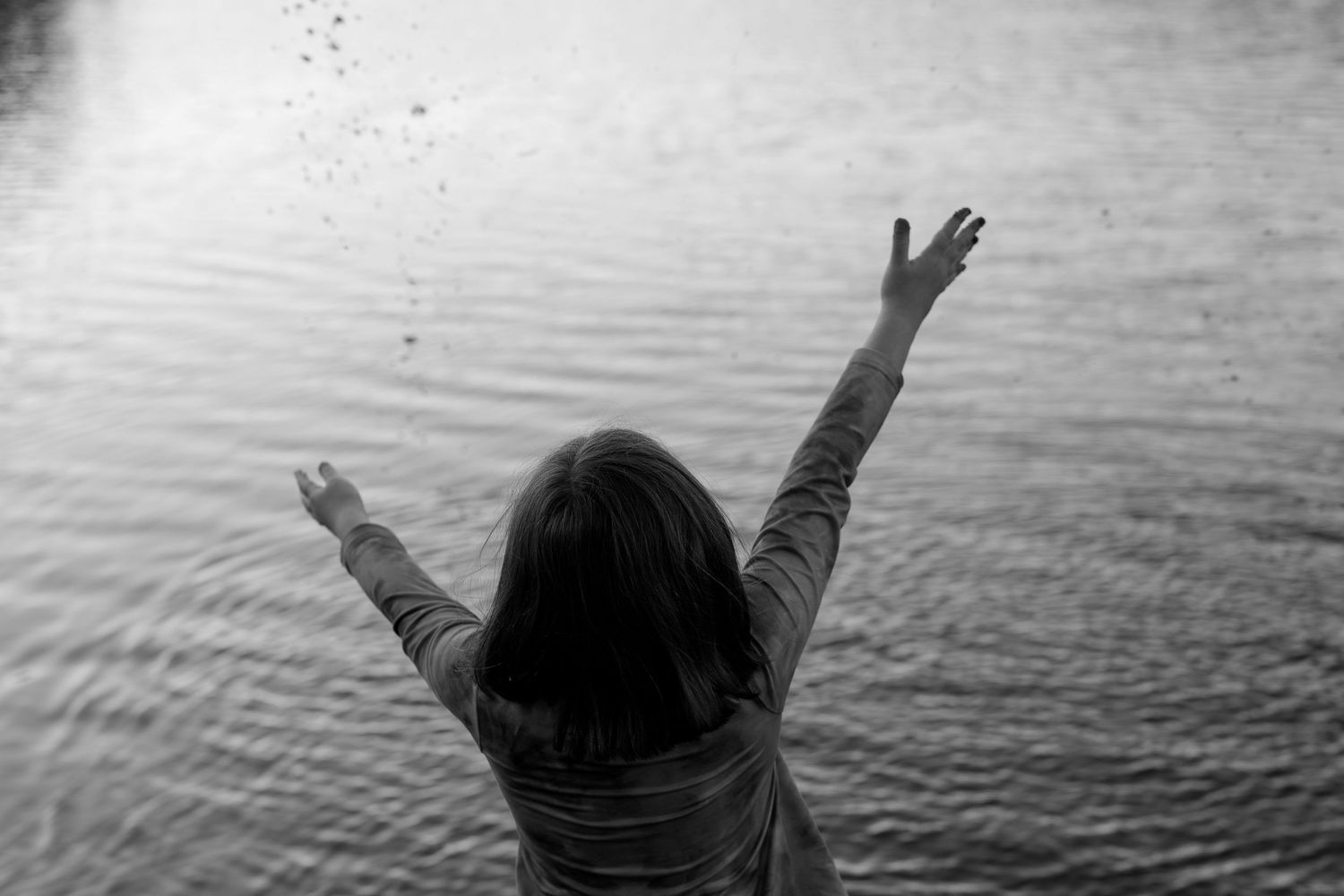 Black-and-white photo of a child throwing pebbles into a lake with arms raised in delight, candid family session