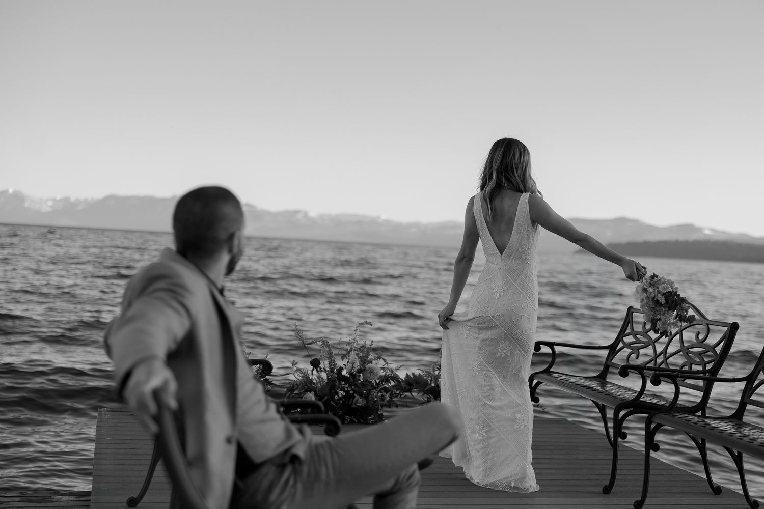 Black-and-white photo of a couple on a lakeside deck with mountains across the water, elegant alpine lake wedding portrait