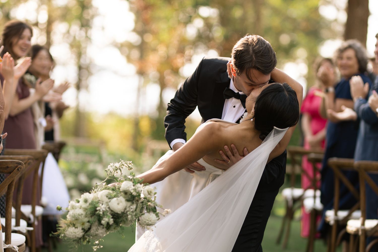 Bride and groom in a dramatic dip kiss surrounded by white florals and cheering guests at an autumn wedding ceremony