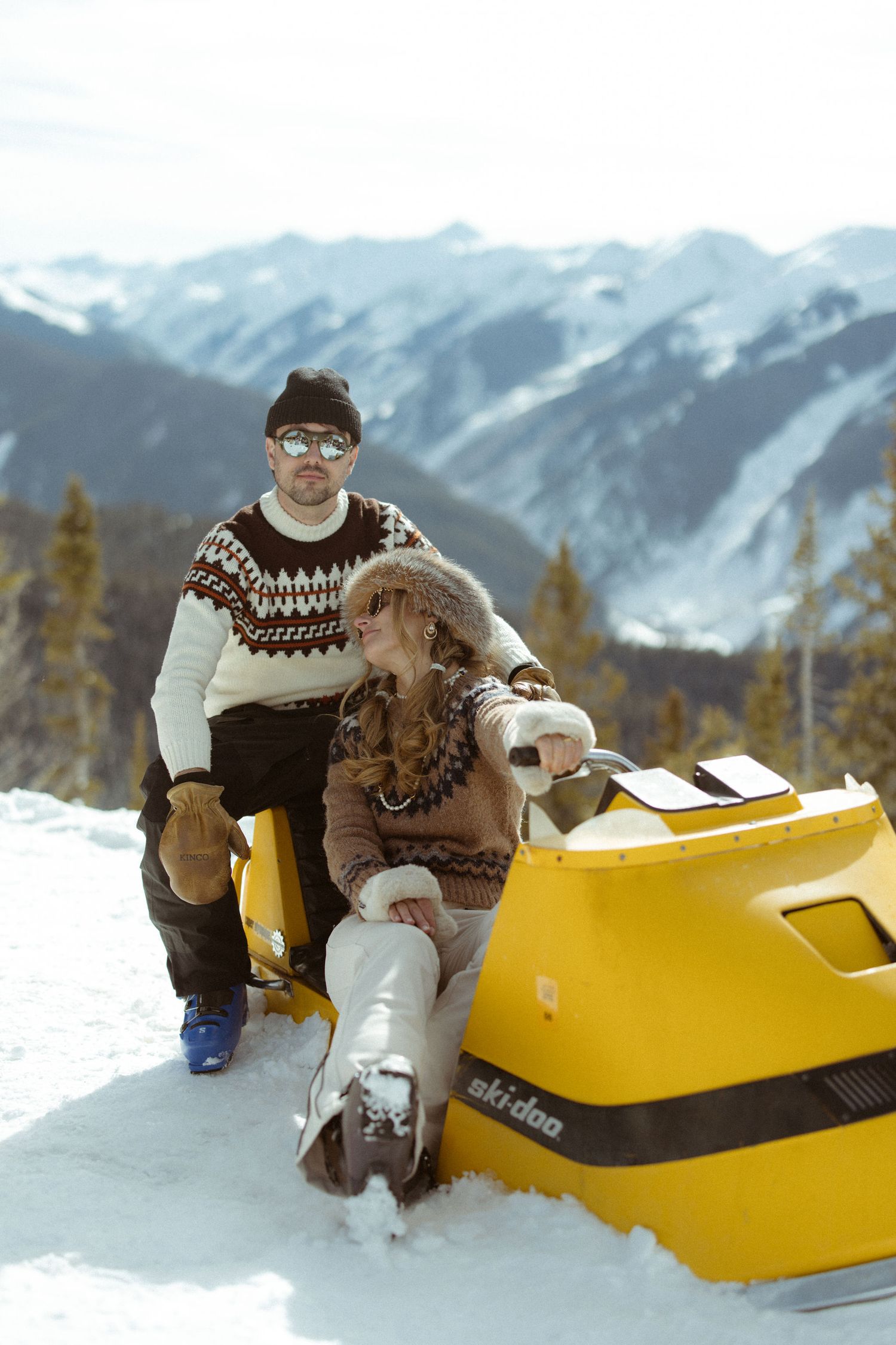Engaged couple on vintage yellow Ski-Doo snowmobile with Aspen ski mountain backdrop during creative engagement session by Sammy Aucella Pho