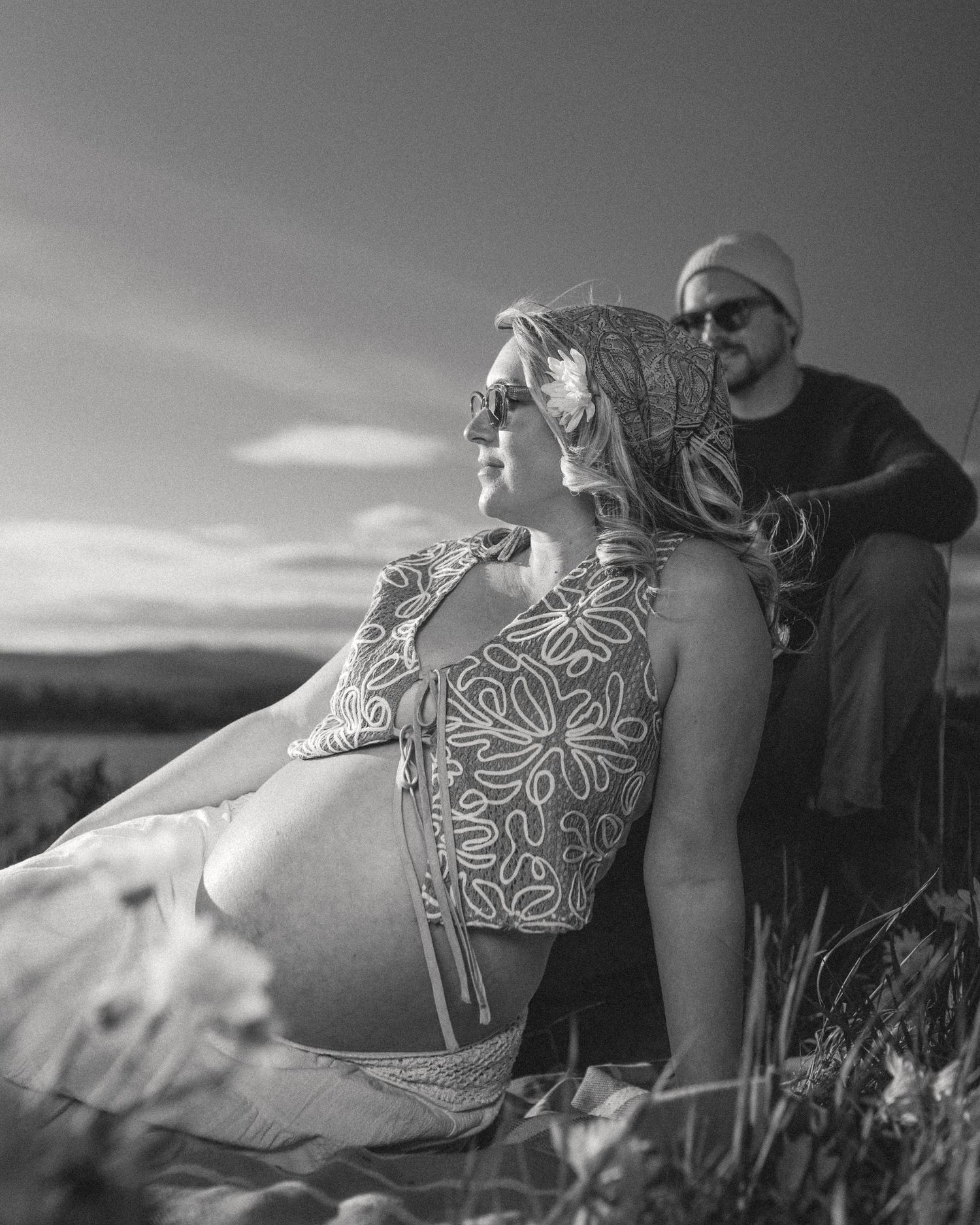 Black-and-white photo of an expectant couple seated in tall grass at sunset, serene outdoor maternity session