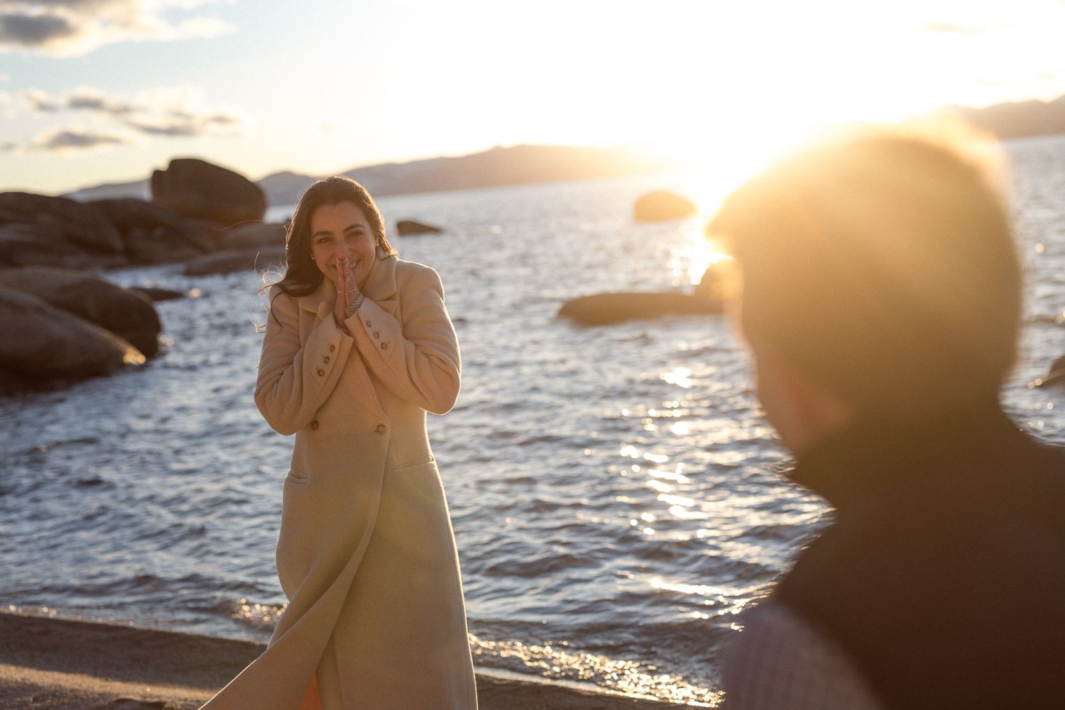 Couple at a sparkling alpine lake at sunset, woman in a cream coat warming her hands, dreamy winter engagement session