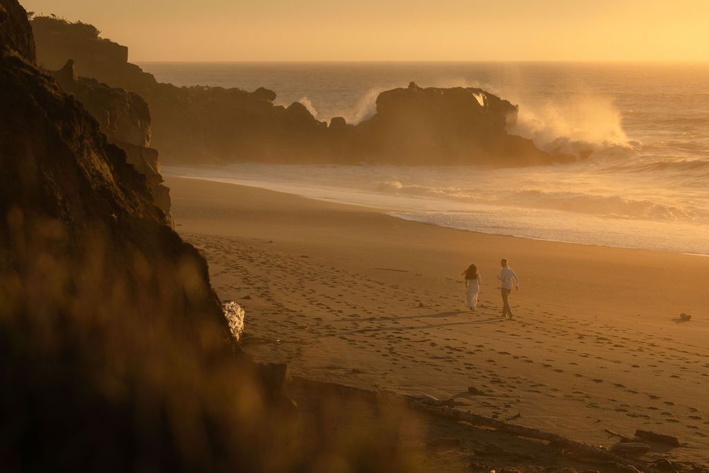 Couple walking a vast golden beach as waves crash against the cliffs, sweeping coastal elopement photography