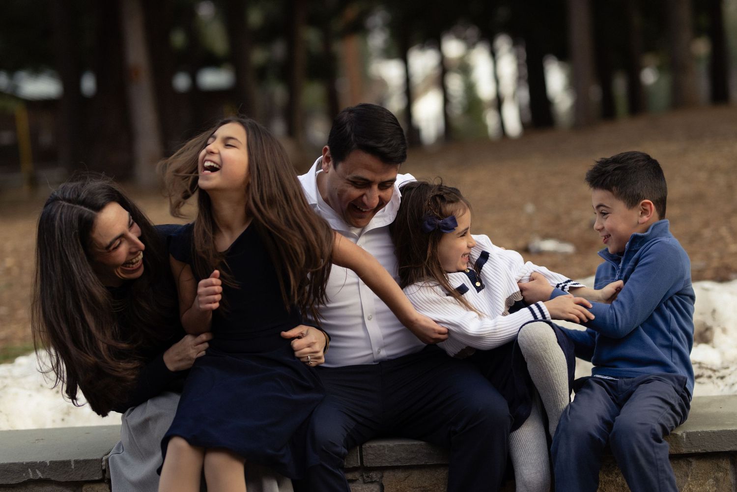 Family of five laughing together on a bench among tall pines, warm and joyful candid family session