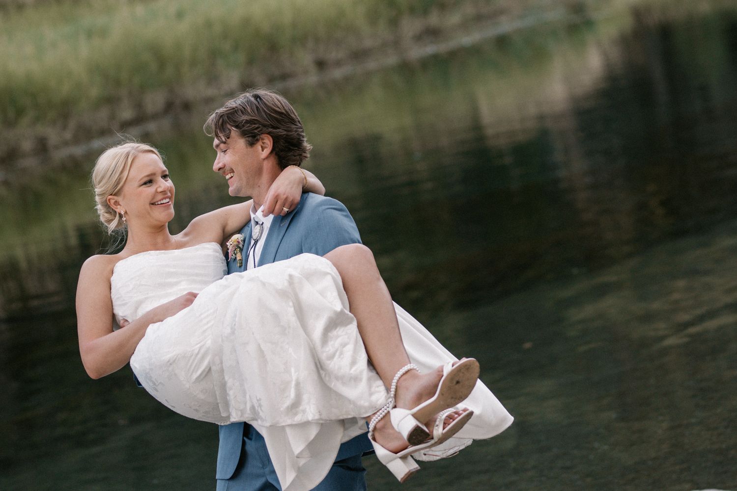 Groom in a blue suit carrying his laughing bride beside a green pond, romantic Spring Ranch Mendocino wedding