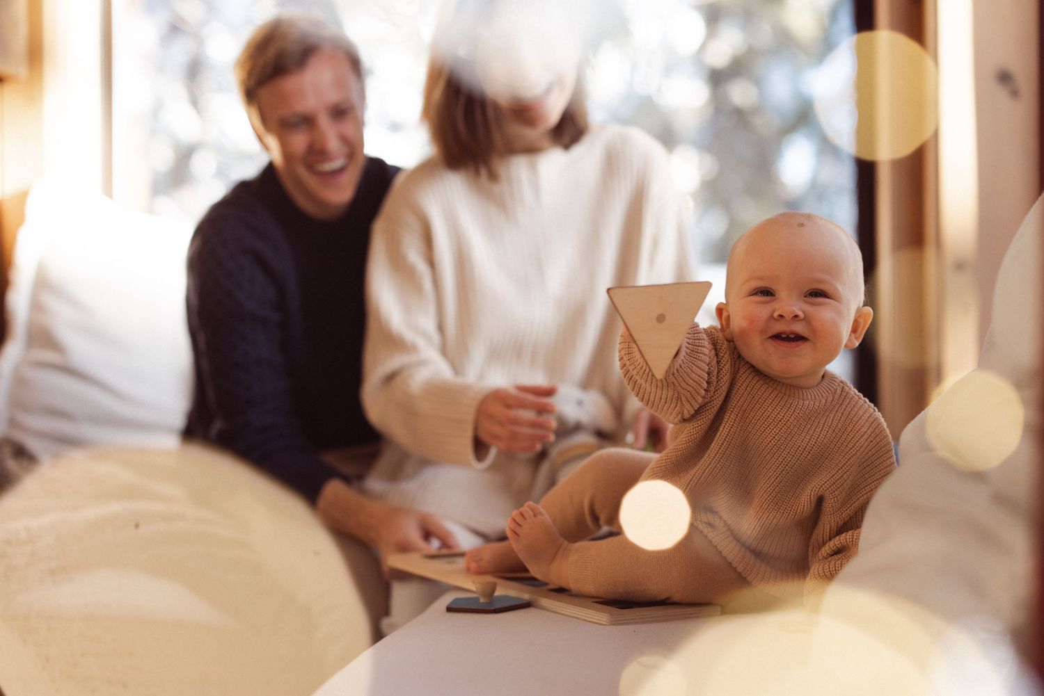 Smiling baby sitting with their parents in a sunlit room full of warm bokeh light, joyful indoor family session