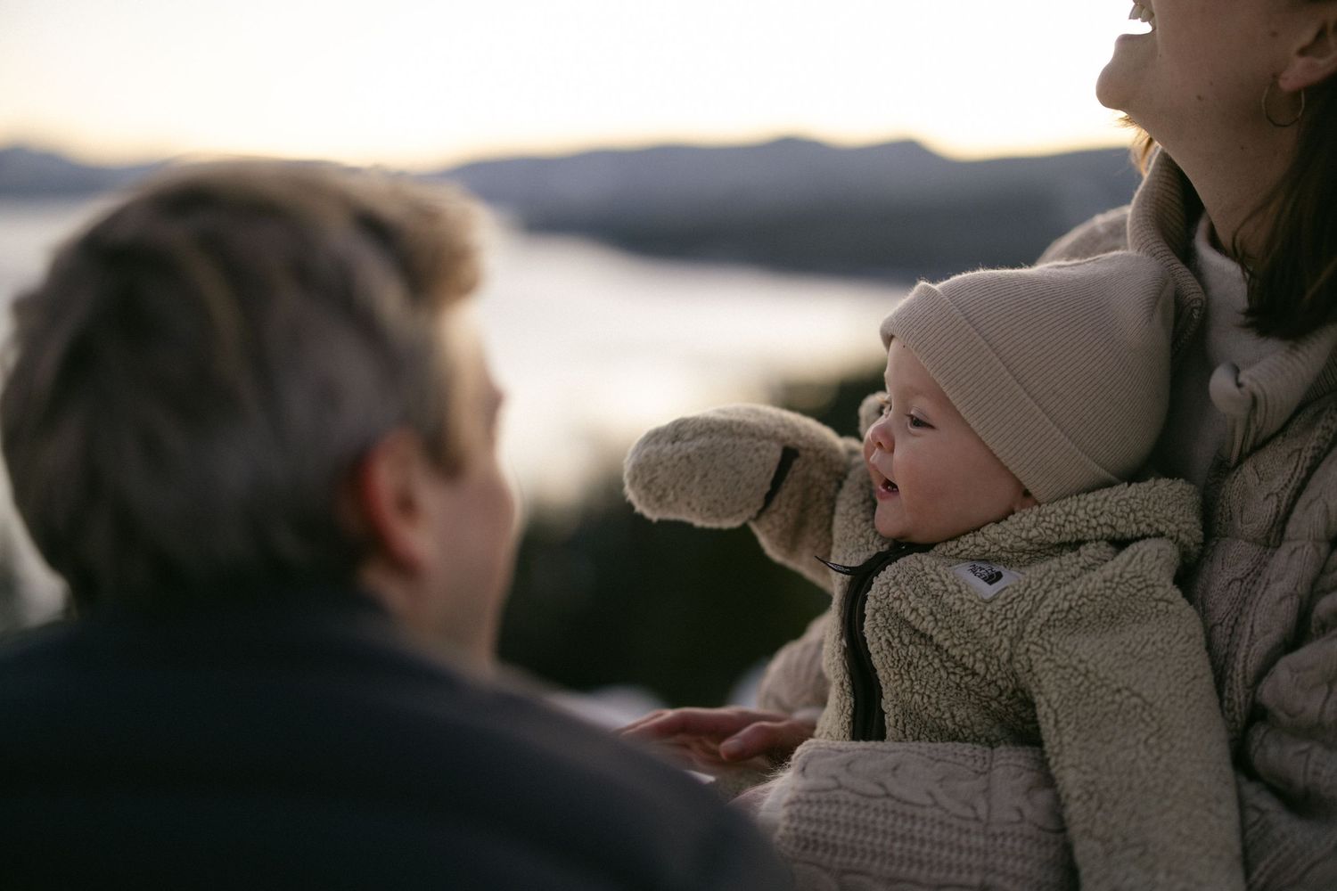 Parents holding their bundled baby beside an alpine lake at golden hour, tender winter family session in Lake Tahoe