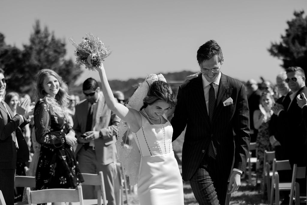 Black-and-white photo of newlyweds recessing down the aisle as the bride raises her bouquet, joyful wedding celebration