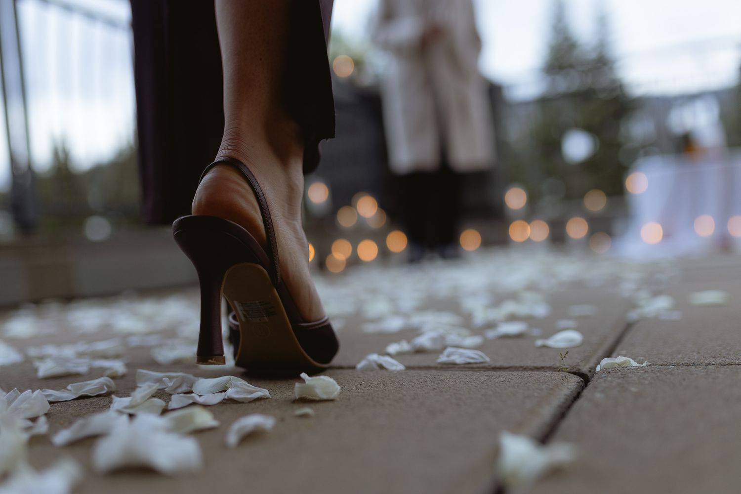 Bride's burgundy heels stepping across scattered petals on a stone path with twinkling lights behind, wedding detail