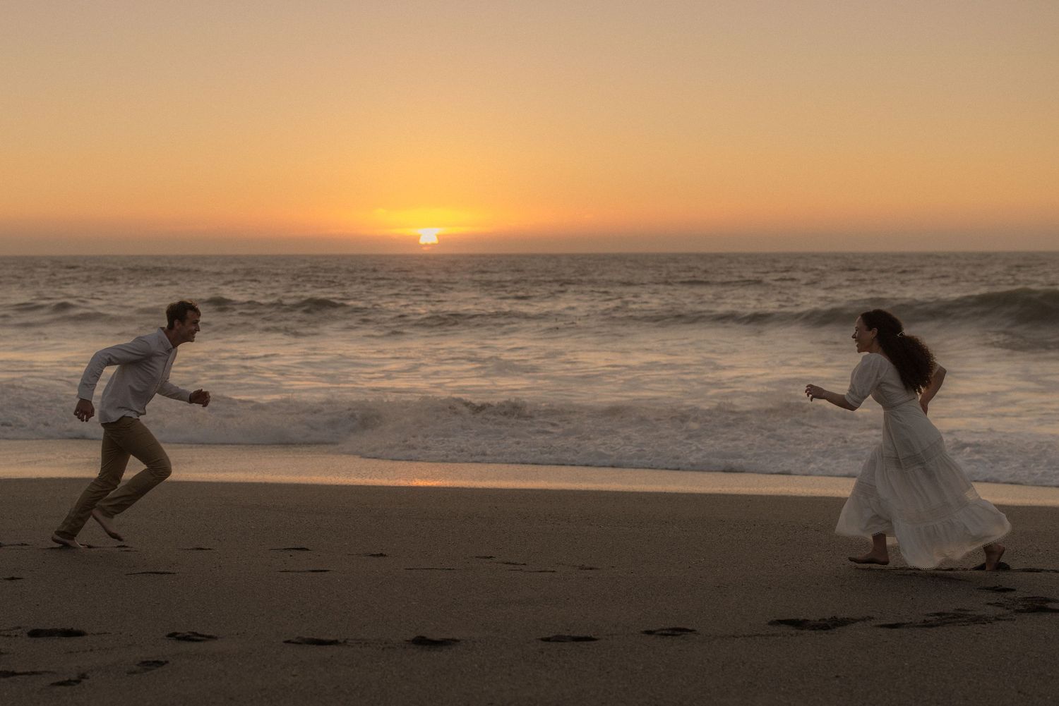 Couple in white playfully chasing each other along the beach as the sun sets over the ocean, joyful engagement session