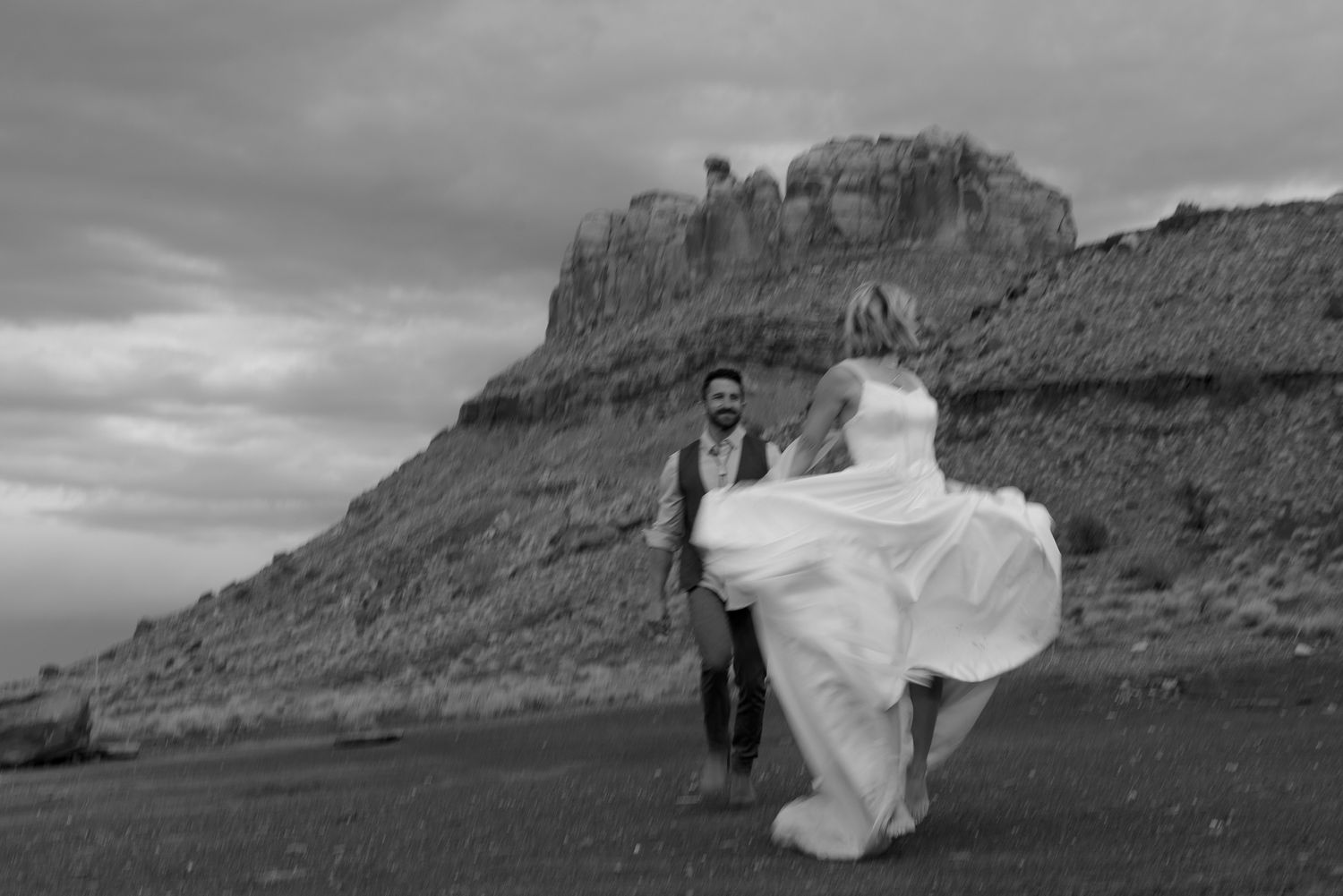 Black-and-white photo of a groom swinging his bride in a billowing gown before desert buttes, adventurous elopement