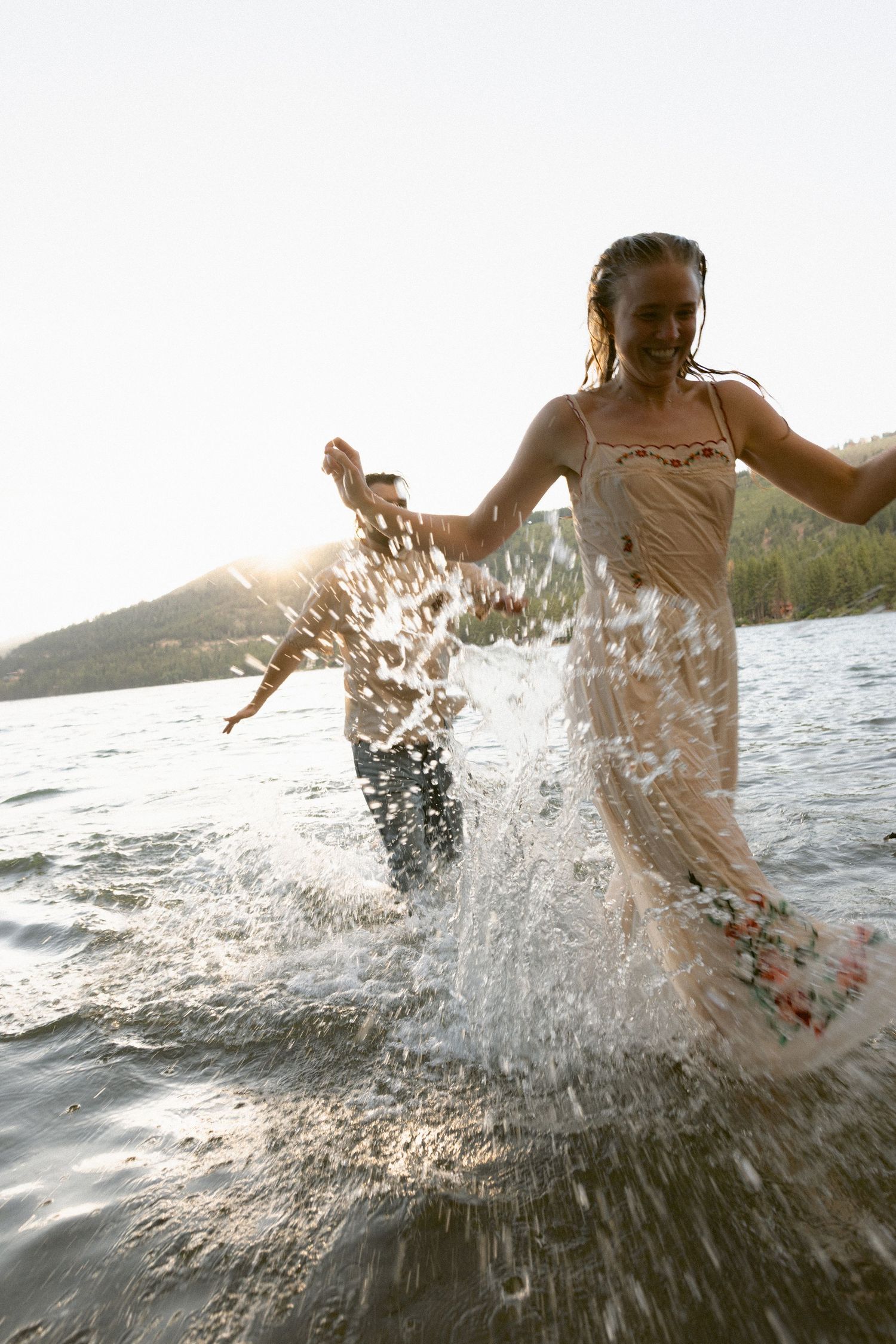 Couple in a flowing gown and suit splashing through a golden-hour mountain lake during a joyful editorial engagement session