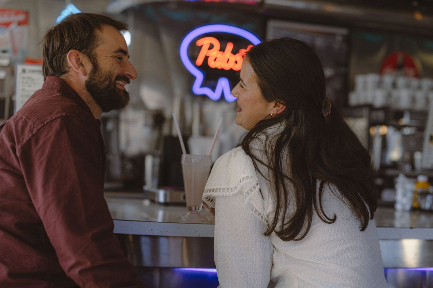 Couple laughing together at a retro bar beneath a glowing neon sign, playful date-night engagement session