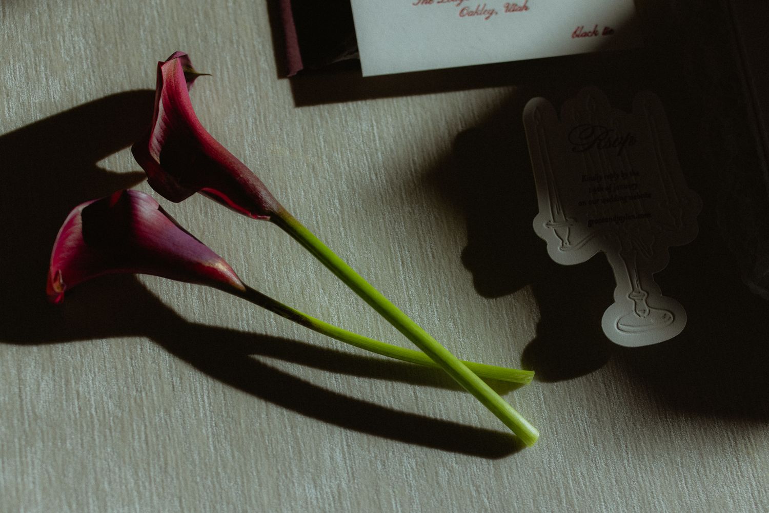 Wedding detail of deep red calla lilies beside the invitation suite casting dramatic shadows, elegant flat lay