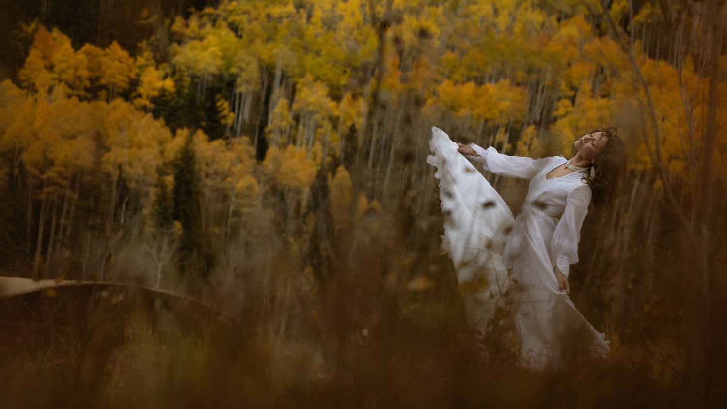 Bride reclining in tall grass amid a golden aspen forest in her white gown, dreamy autumn Aspen Colorado bridal portrait