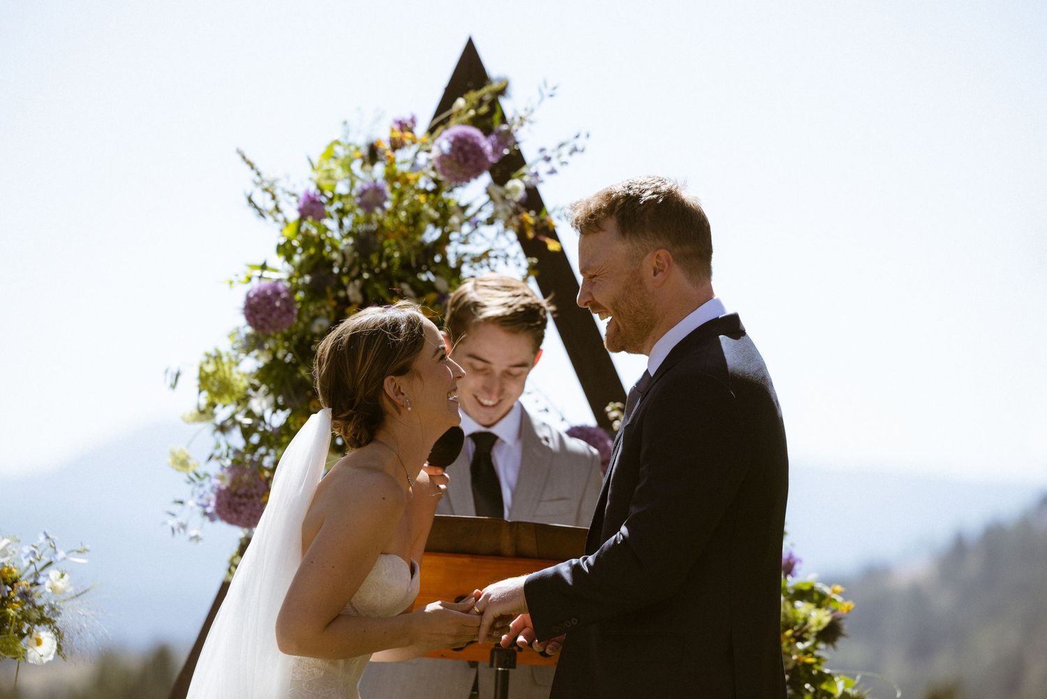 Bride and groom exchanging vows at a triangular wildflower arch beside an alpine lake, sunlit mountain wedding ceremony