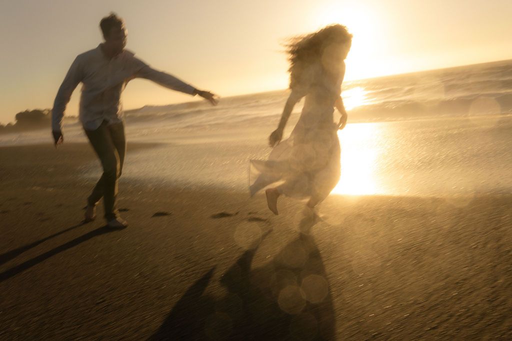 Couple playing and chasing each other across the sand at sunset, joyful coastal couples session