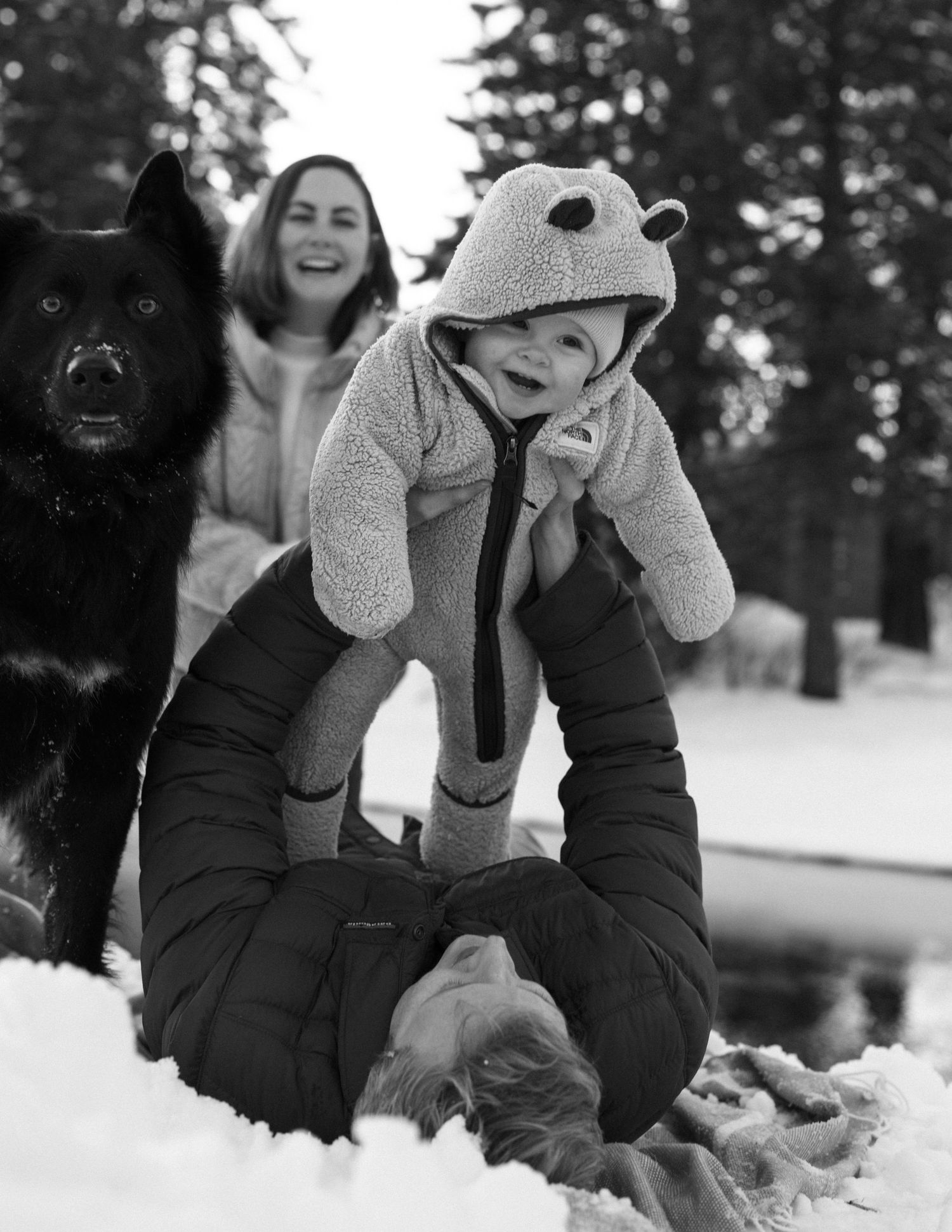 Black and white family portrait in the snow with their dog and toddler in a bear suit, playful winter family session