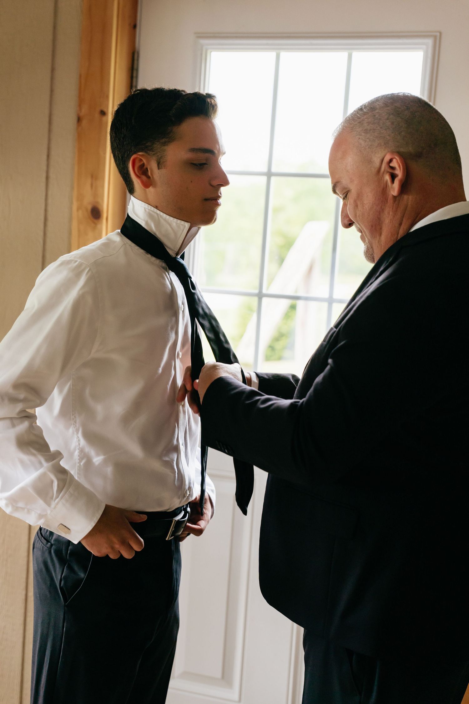 Two people in formal attire stand by a window as one helps adjust the other's tie and clothing.