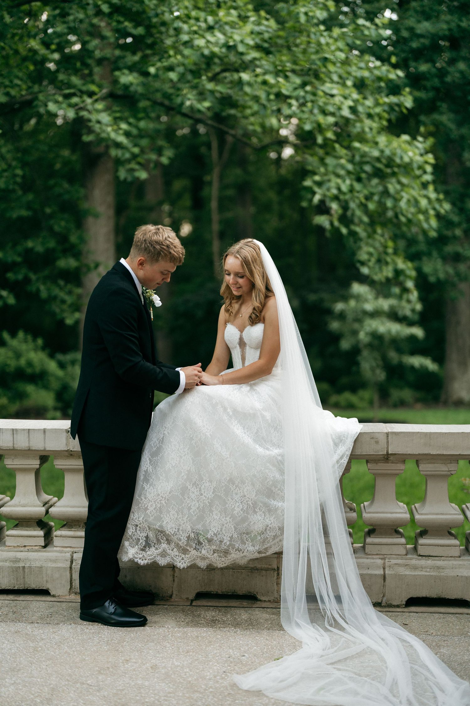Bride in lace gown and veil sits on stone balustrade as groom in black tuxedo holds her hands in lush garden.
