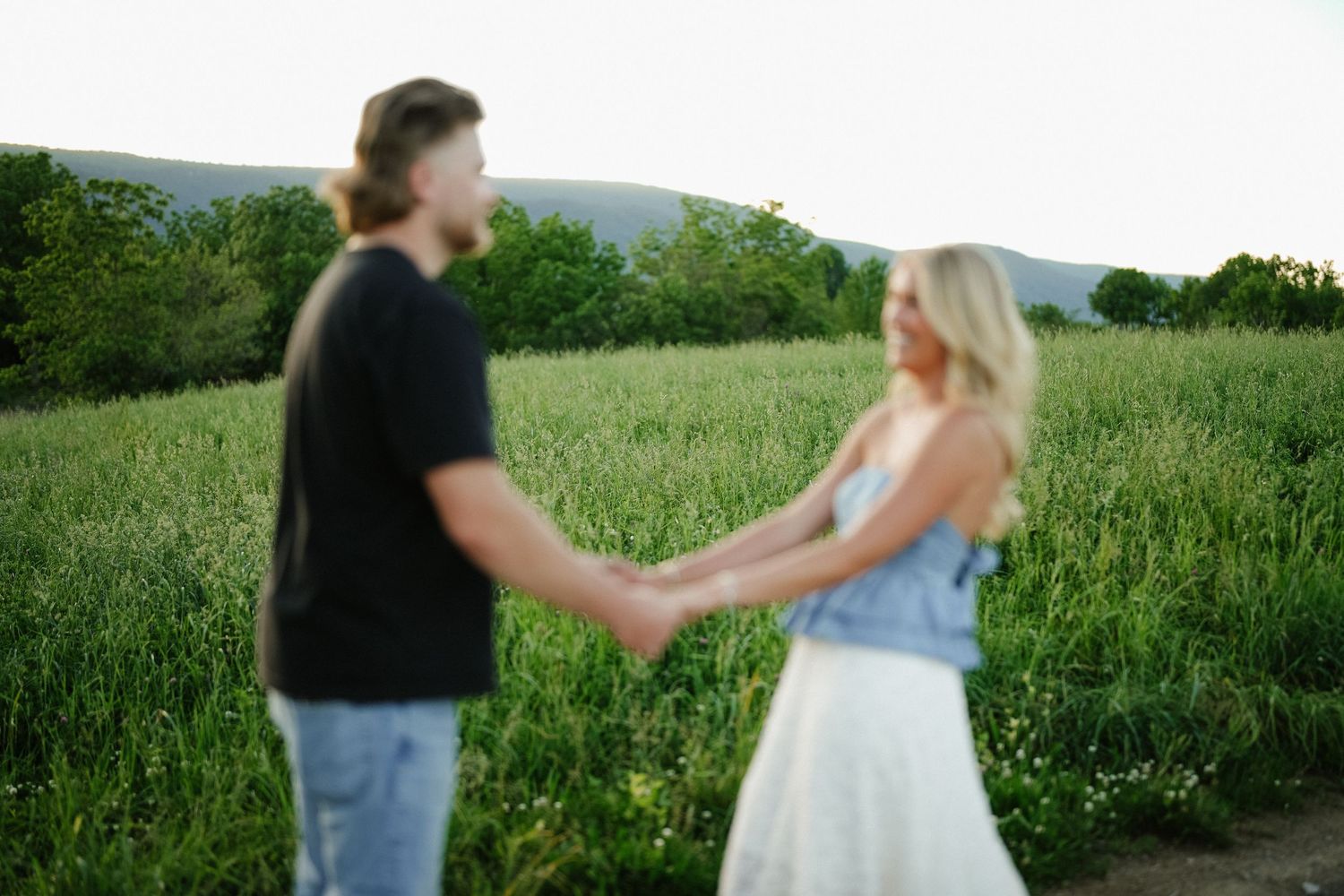 Couple holding hands in a lush green field at sunset, man in black shirt and woman in white skirt.