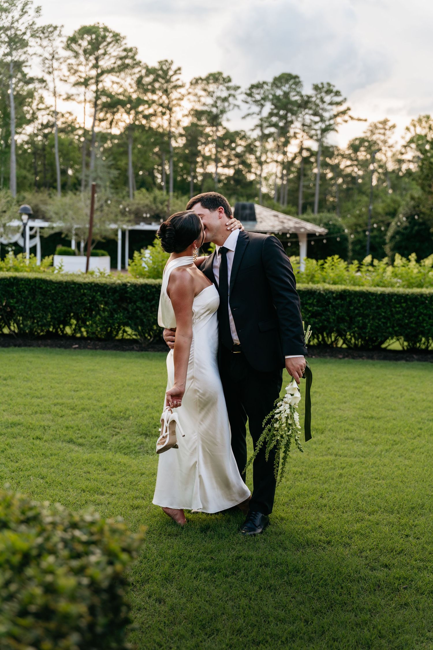 Bride and groom share a kiss on lush green lawn at golden hour, surrounded by manicured hedges and tall pine trees.