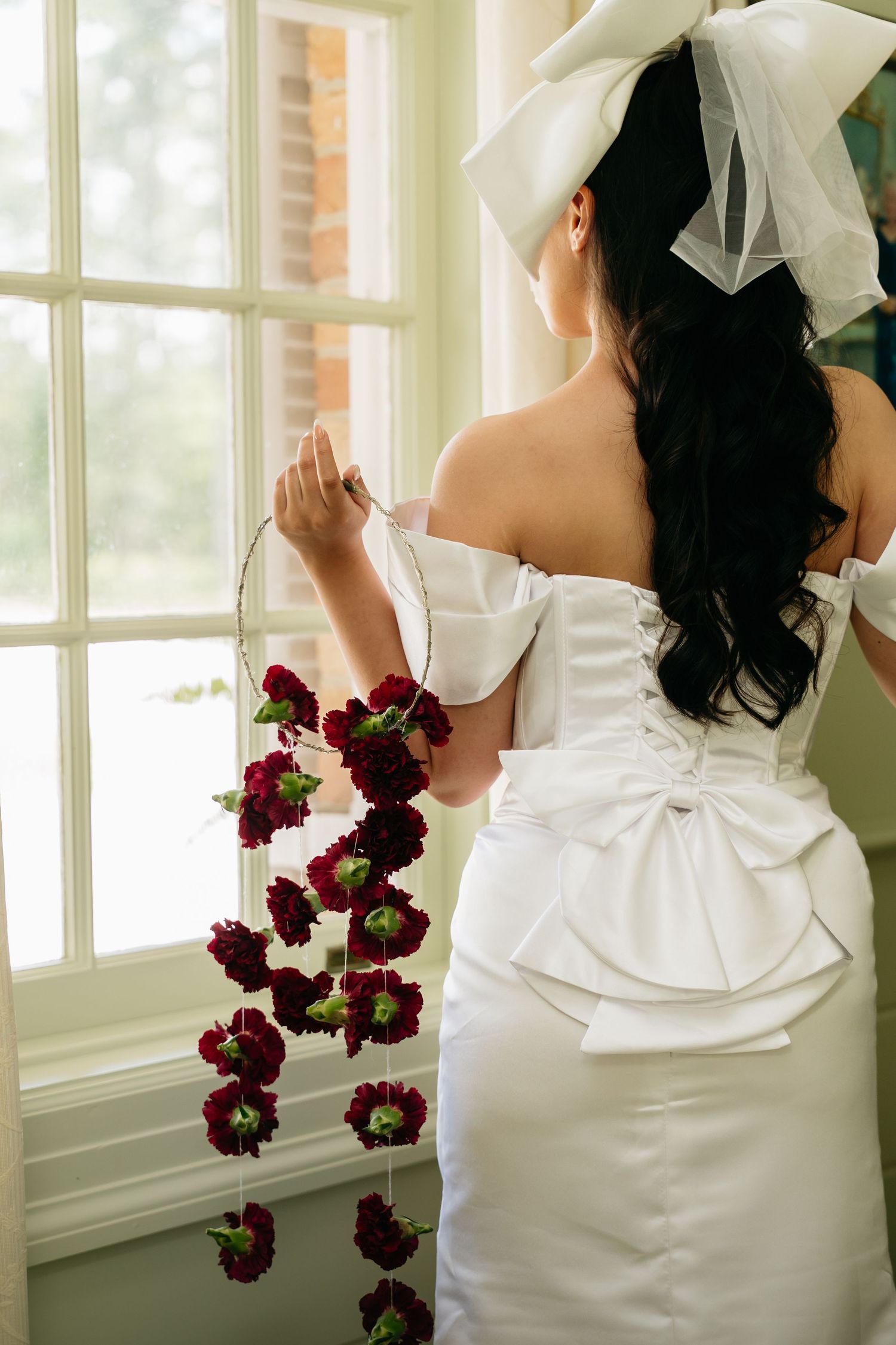 Bride in white off-shoulder gown with bow detail holds burgundy floral garland while standing at a window.