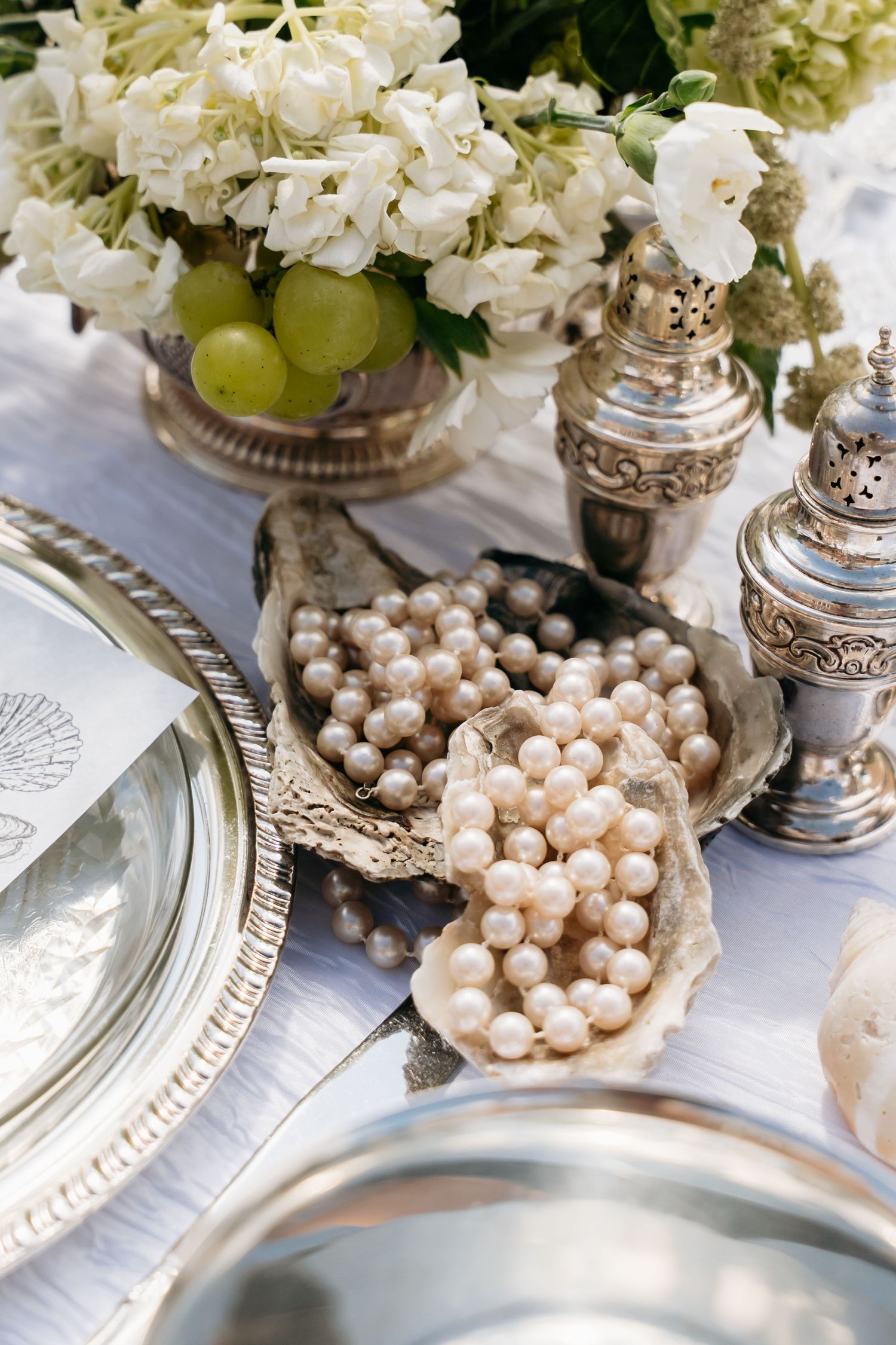 Elegant table setting with pearl necklaces in oyster shells, silver salt shakers, and white floral arrangement with green grapes.