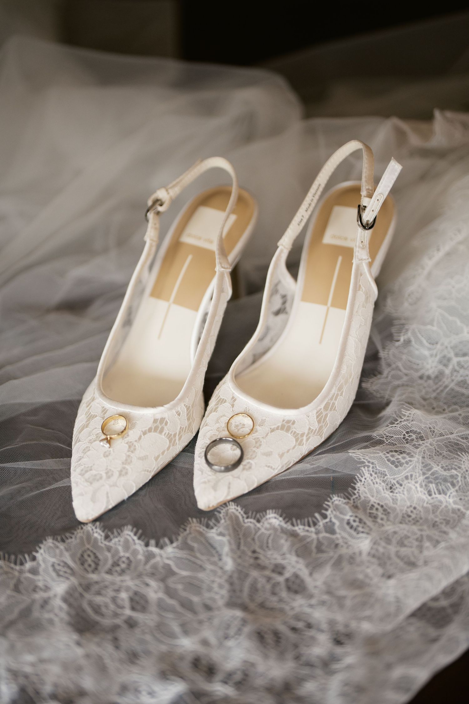 White lace slingback wedding heels with gold rings resting on top, displayed on a delicate lace veil.