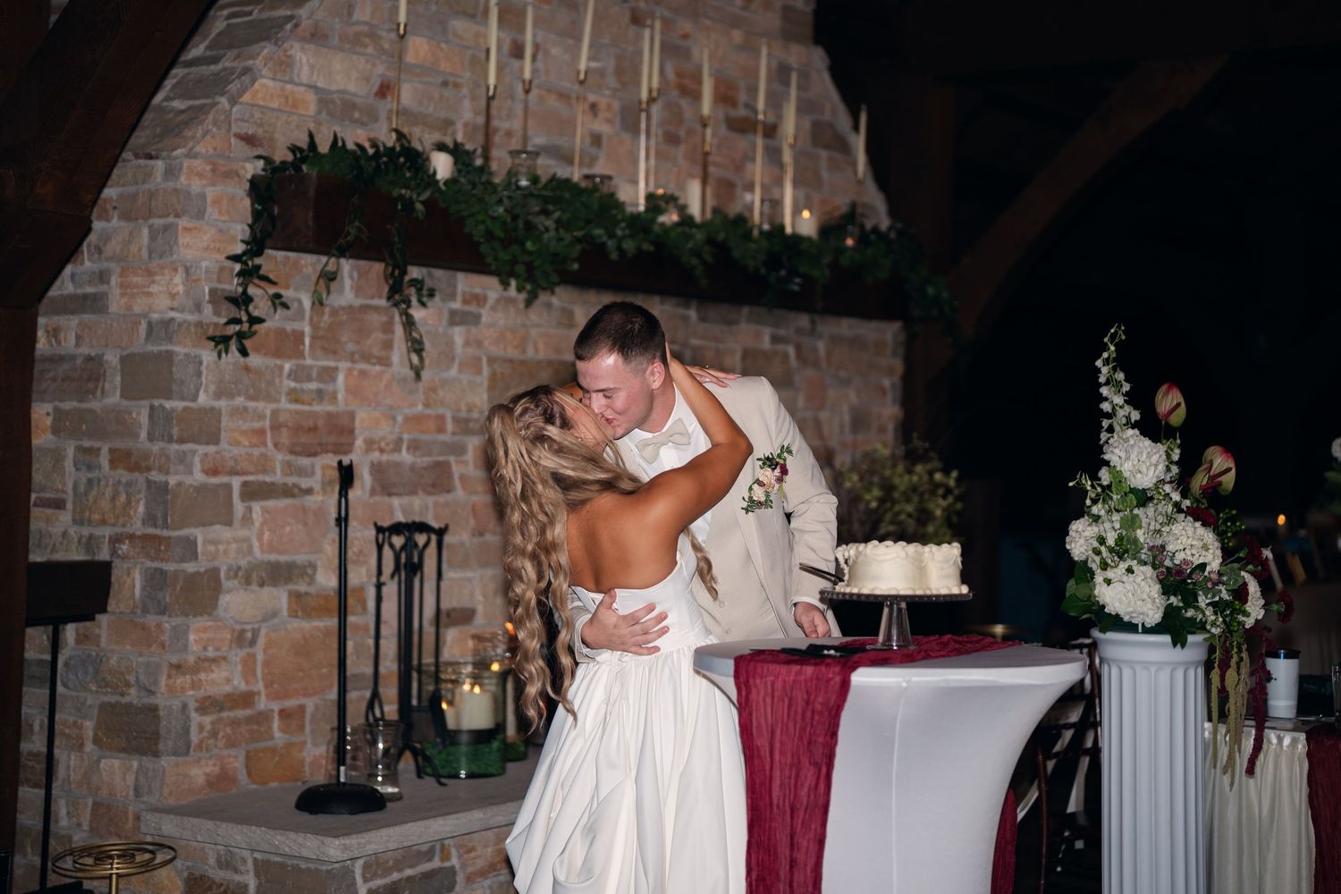 Bride and groom share a romantic kiss near a rustic brick fireplace beside a white wedding cake at their reception.