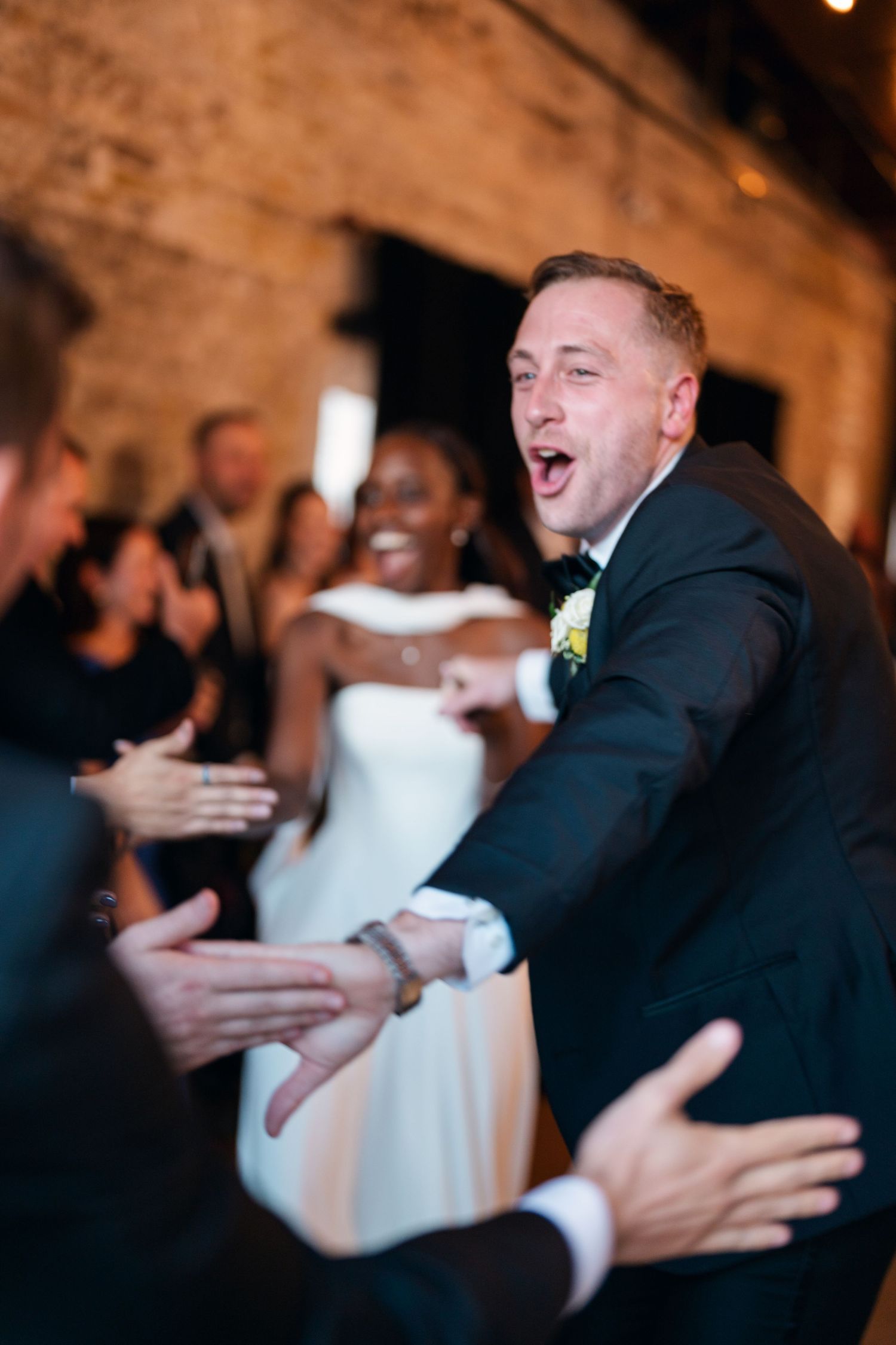 Joyful groom in tuxedo high-fiving guests while bride in white gown smiles during wedding reception entrance.