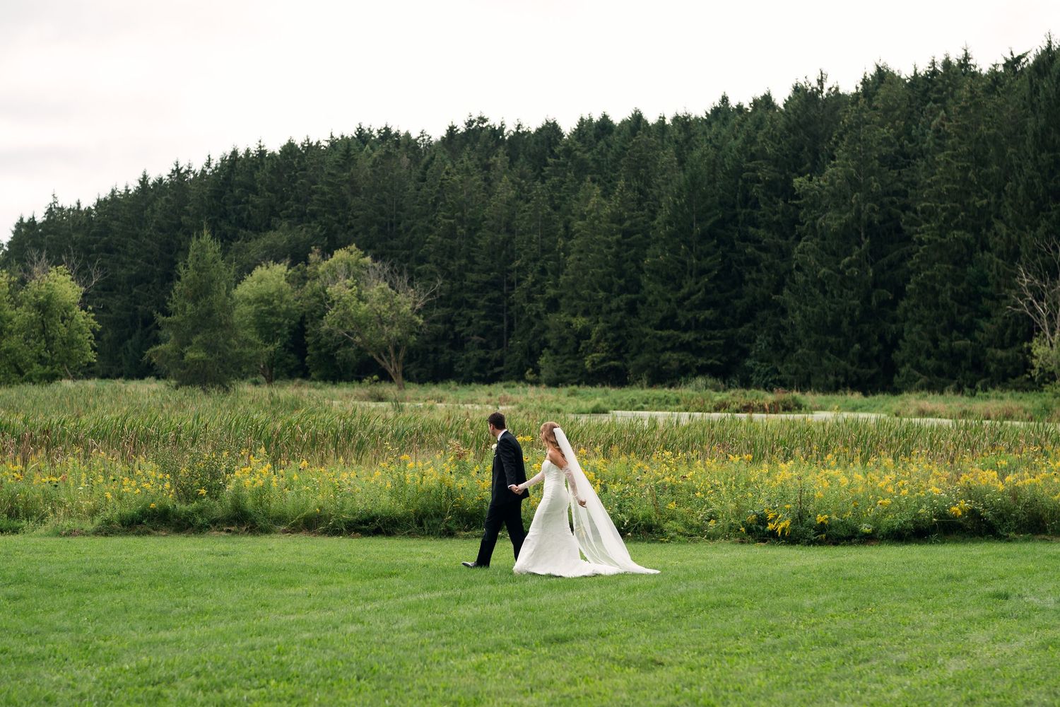 Bride and groom walking hand in hand across a lush green meadow with wildflowers and a dense forest backdrop.