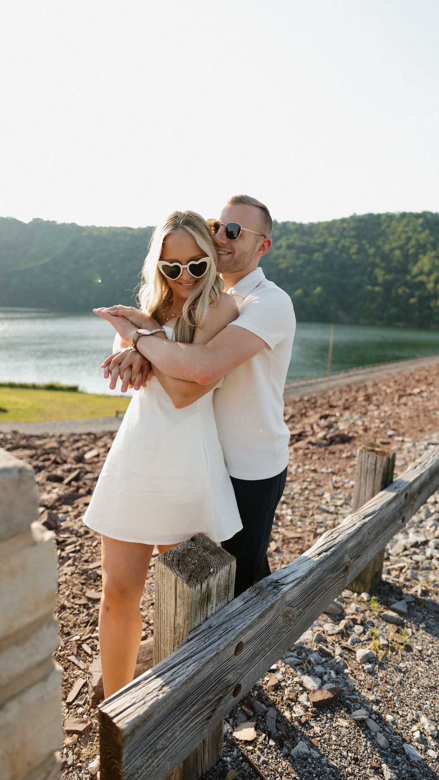 A couple posing together on a rocky shoreline near a lake wearing sunglasses.