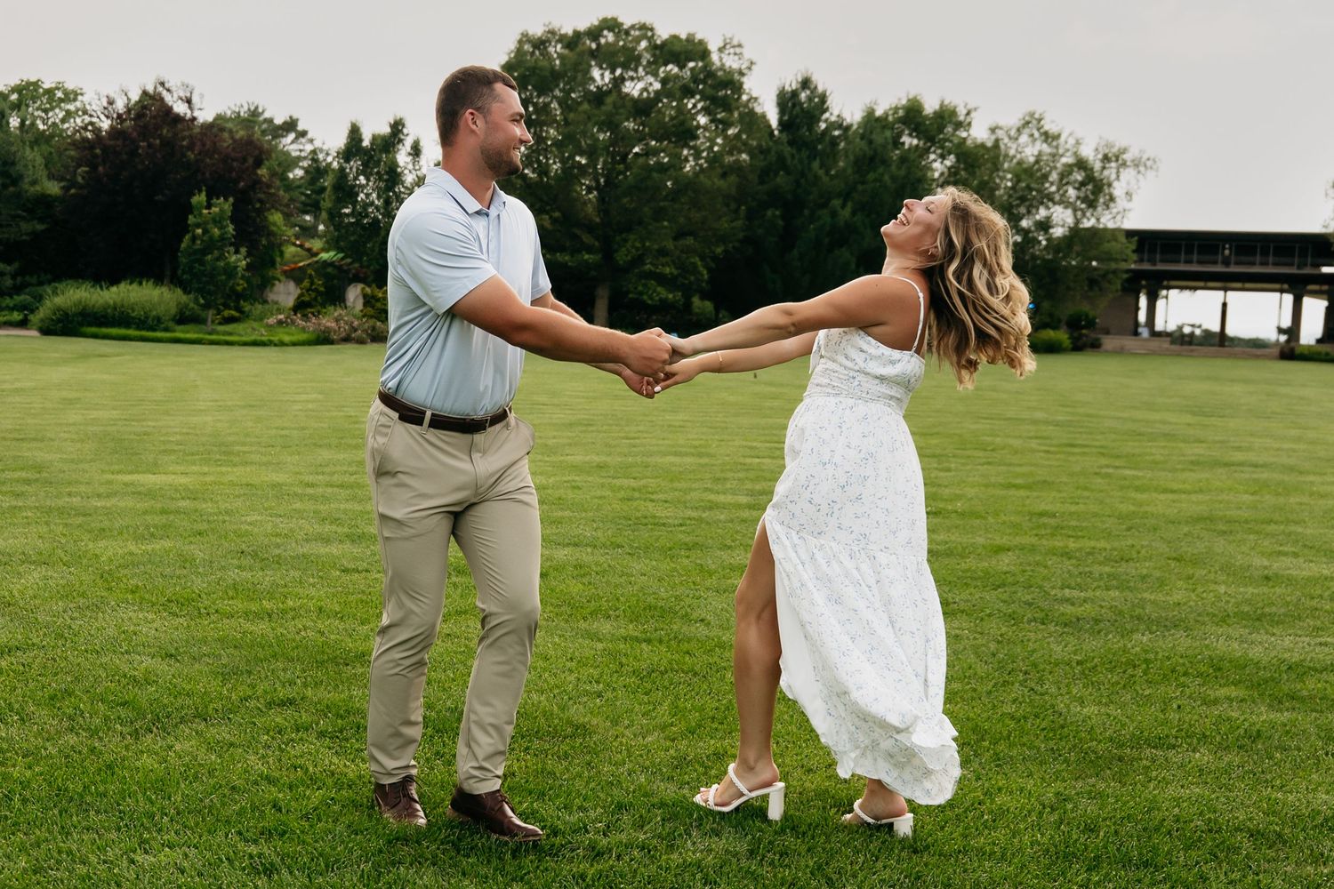 Couple in white and khaki outfits dancing playfully on a lush green lawn during a sunny day.