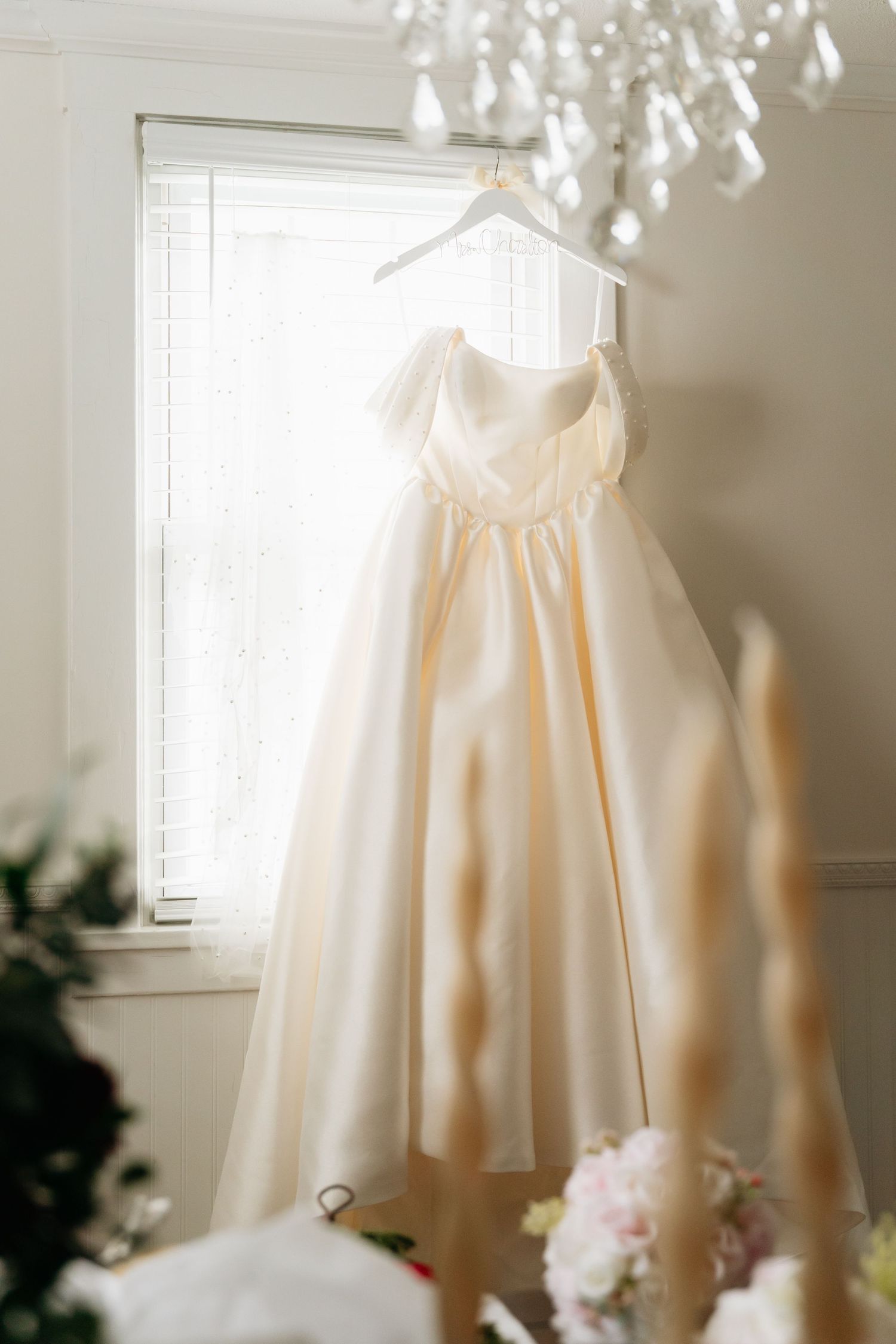 A delicate cream-colored wedding dress hangs elegantly by a sunlit window with soft lighting creating a romantic atmosphere.