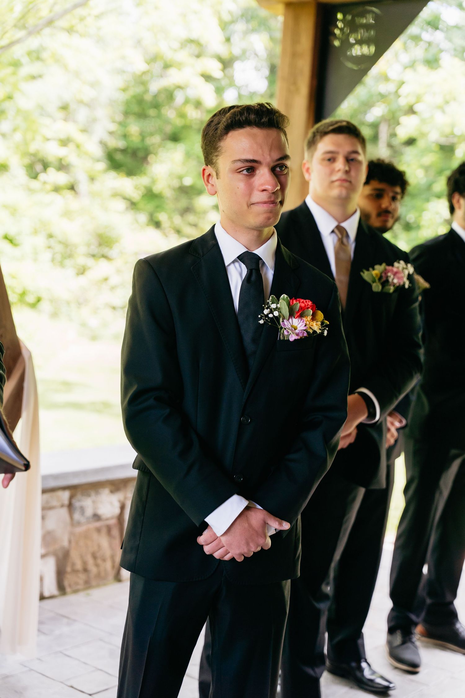Groomsmen in dark suits with boutonnieres stand at attention during an outdoor wedding ceremony.