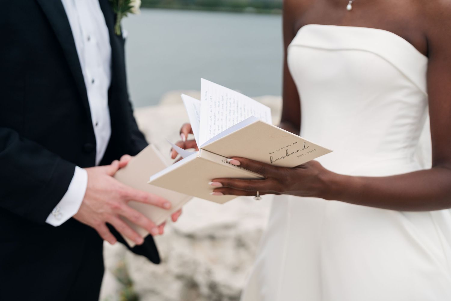 Bride and groom exchanging wedding vows, holding elegant booklets during an outdoor waterfront ceremony.