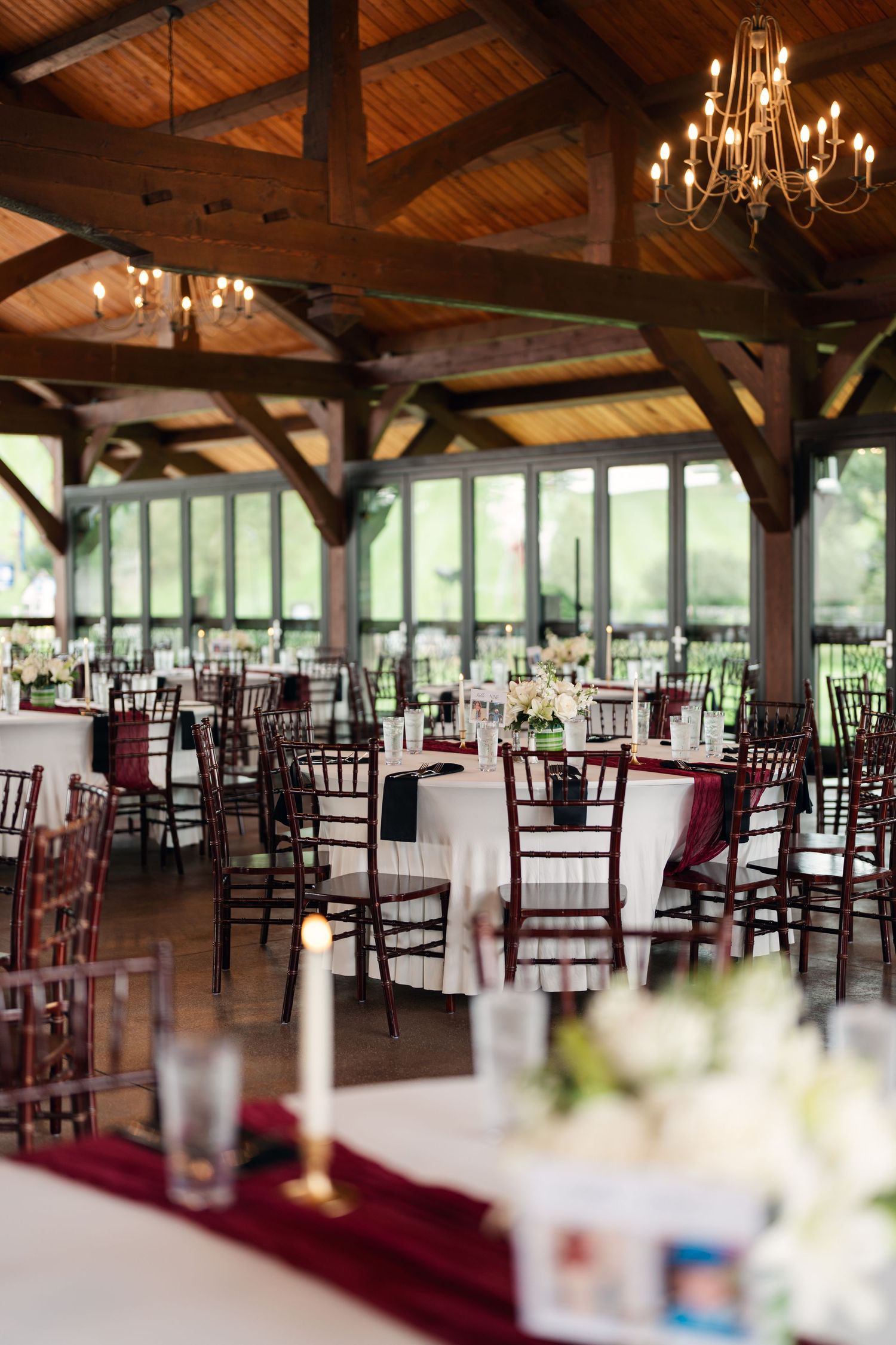 Elegant wedding reception venue with wooden beamed ceiling, chandeliers, and burgundy table runners.
