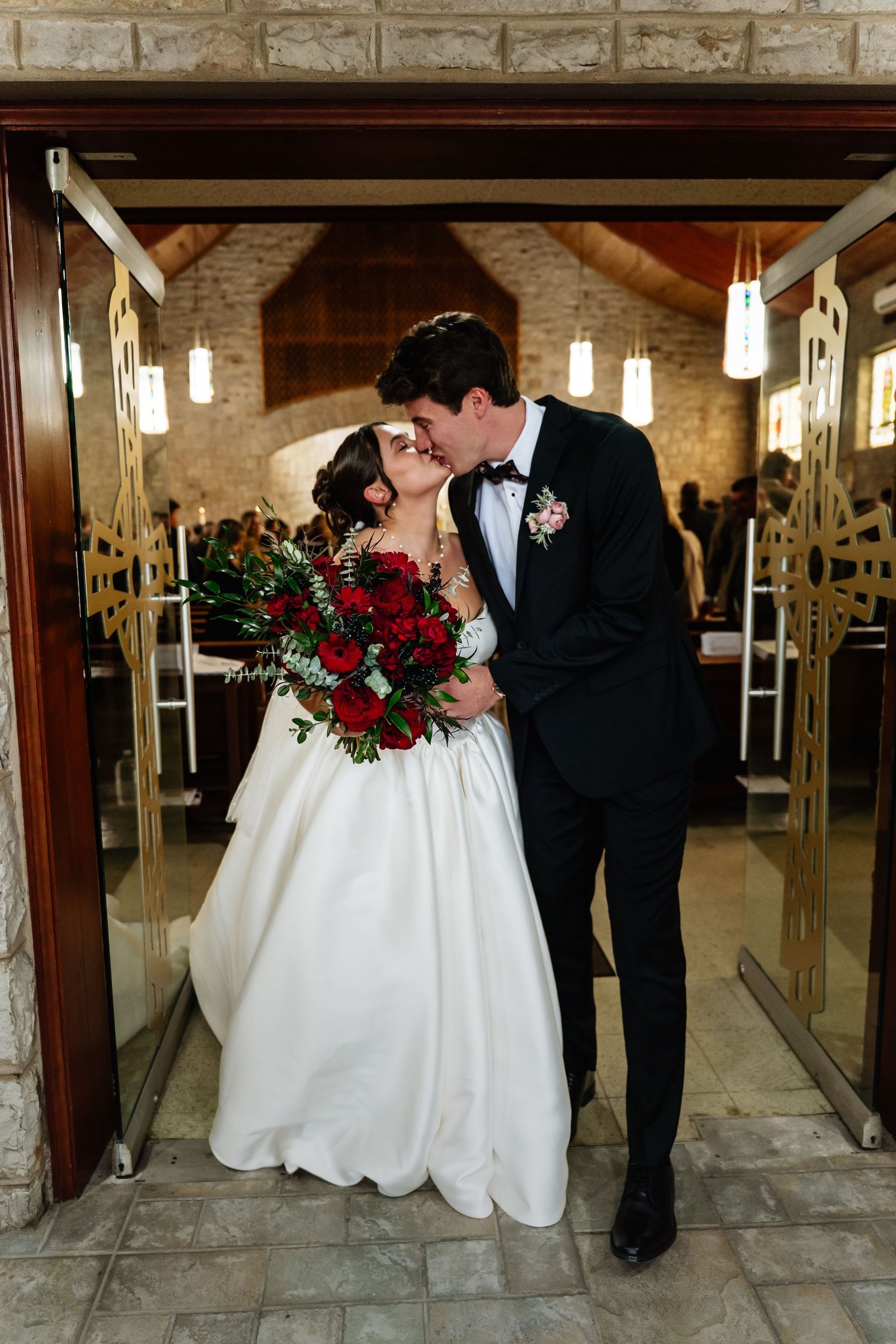 Elegant wedding couple sharing an intimate moment in a stone-walled venue with hanging lights and a red rose bouquet.
